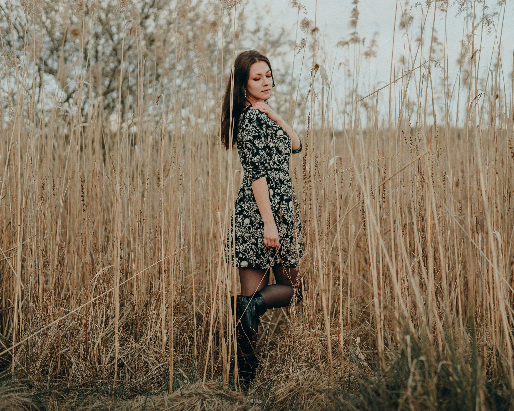 Eine junge Frau in einem schwarzen Kleid mit weißen Mustern steht in einem Feld mit vertrocknetem Gras und trägt schwarze Stiefel.
