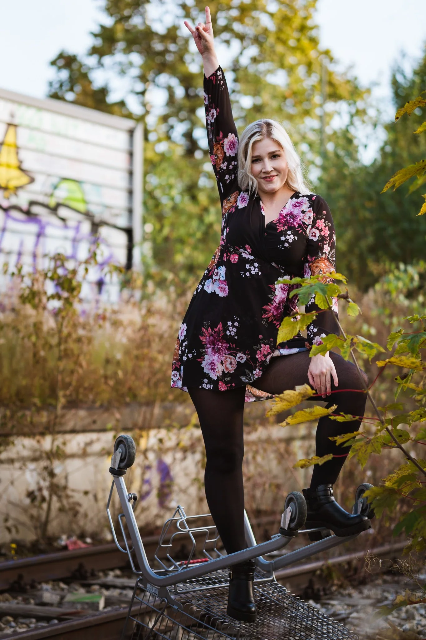Junge Frau trägt schwarzes Kleid mit rosa und lila Blumen, steht auf Einkaufswagen auf Bahngleisen im Freien, umgeben von Büschen und Bäumen.