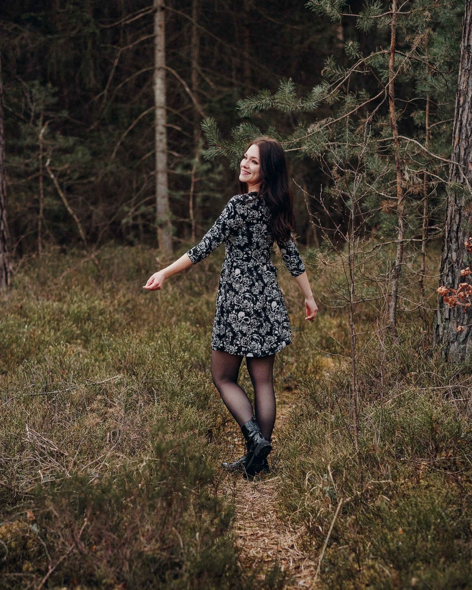 Eine lachende Frau in einem schwarzen Kleid mit weißen Blumenmustern, schwarzen Strumpfhosen und Stiefeln tanzt in einem Wald mit Bäumen und niedrigem Gebüsch.