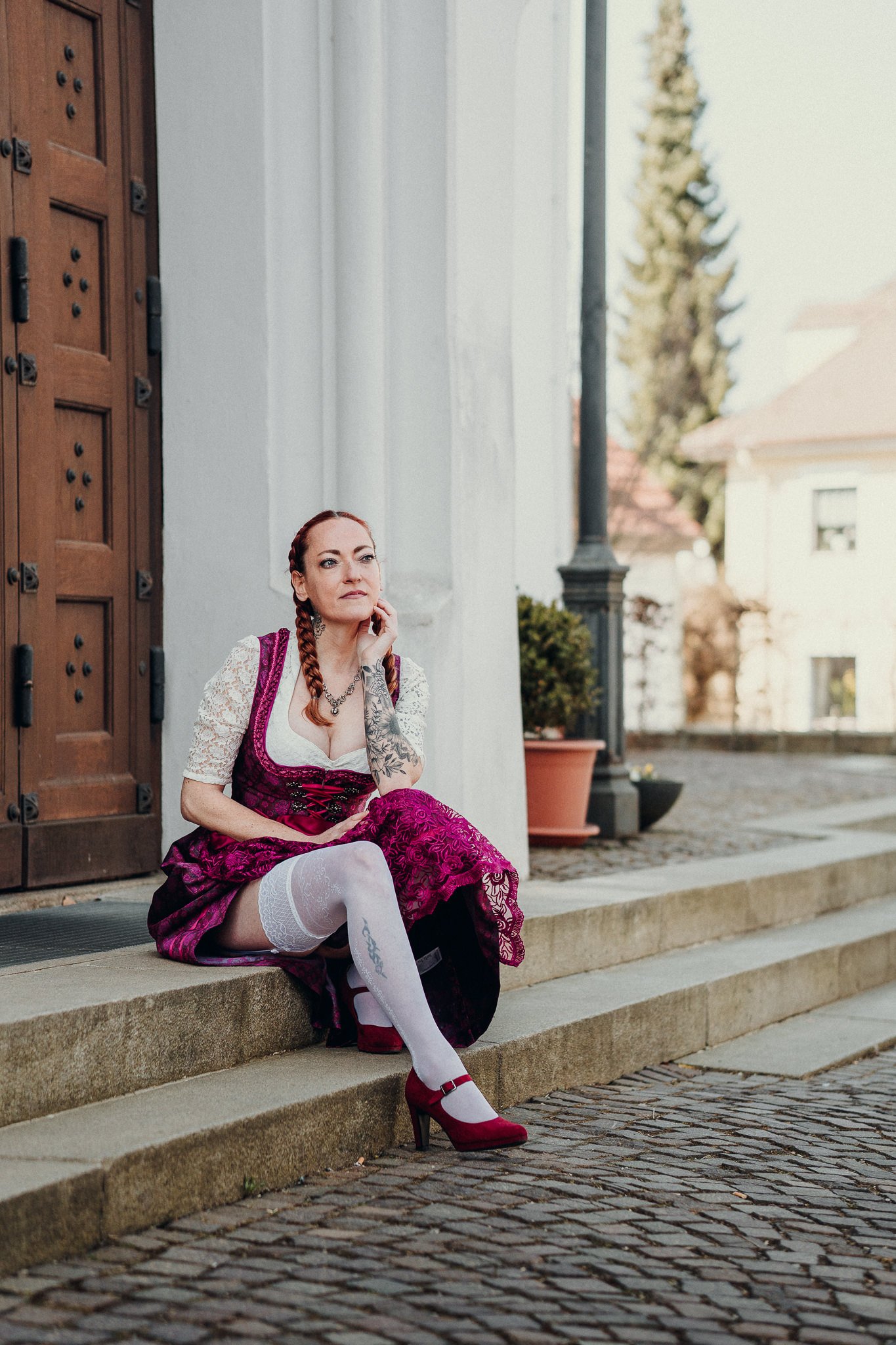 Eine Frau im traditionellen Dirndl sitzt auf den Stufen vor einer Tür mit einem nachdenklichen Gesichtsausdruck.