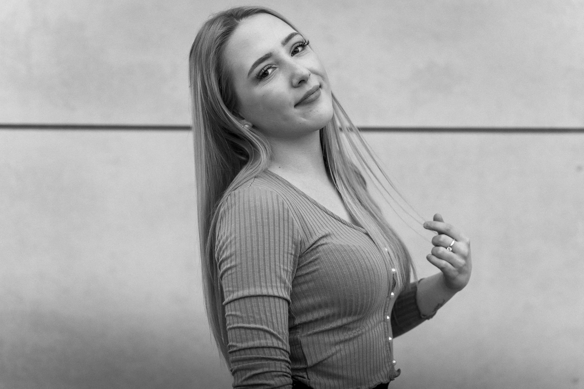 Junge Frau mit langem Haar, lächelnd, in einem Rippstoff-Oberteil, steht vor einer glatten Wand, die in zwei Hälften geteilt ist.