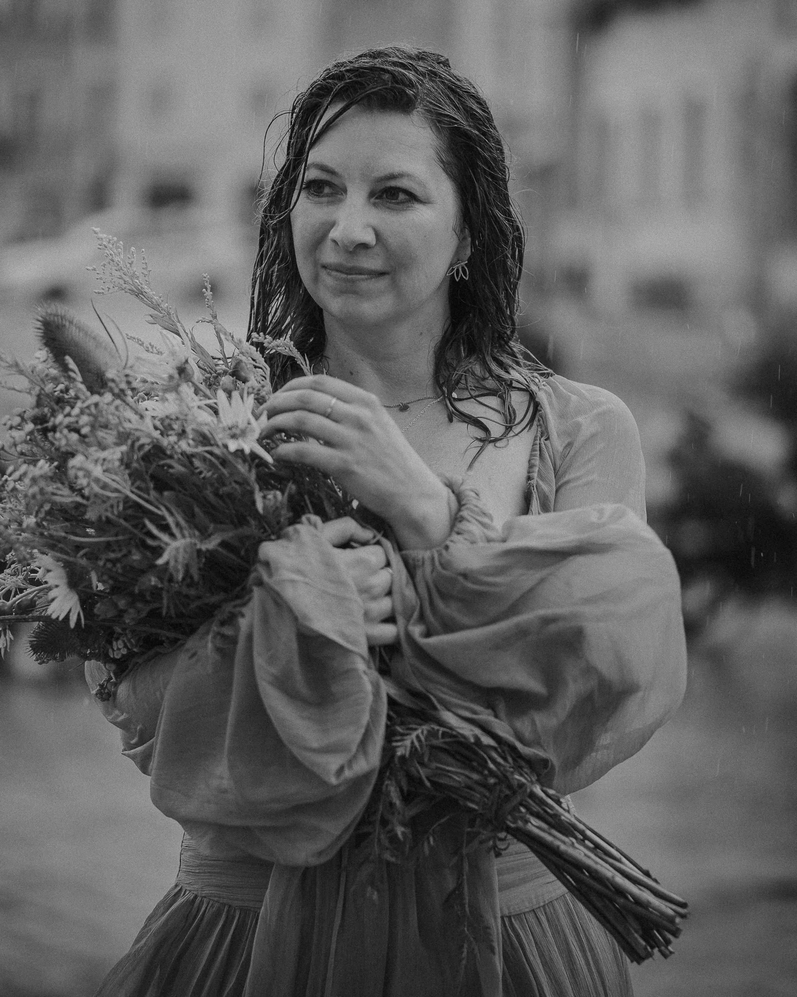 Eine Frau mit nassem Haar hält einen Blumenstrauß und schaut nach rechts, im Hintergrund regnet es.