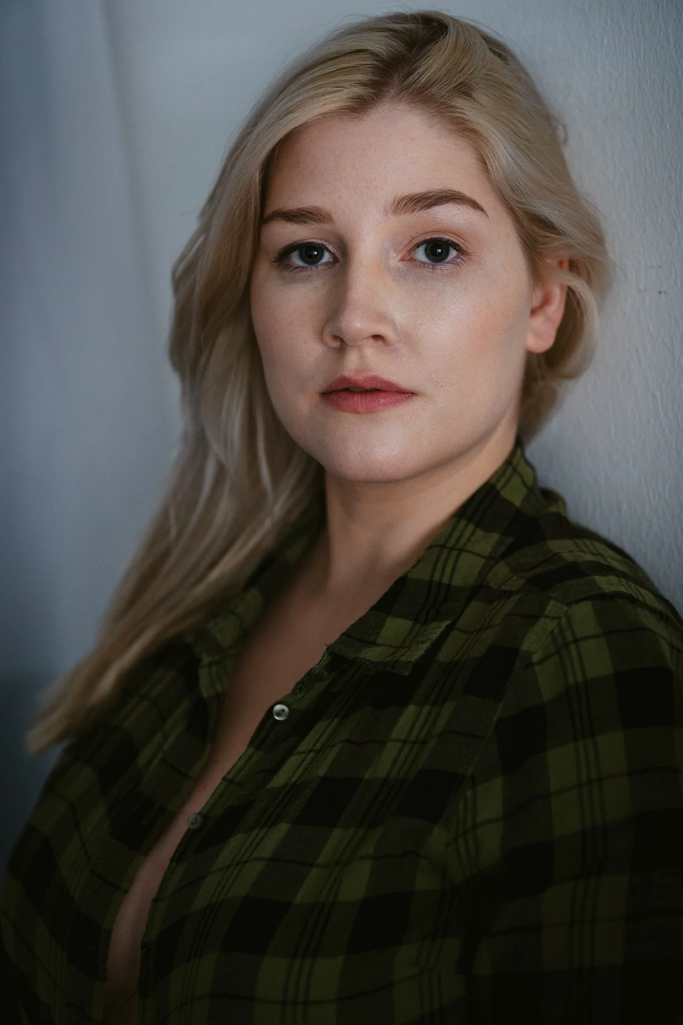 Nahaufnahme einer jungen Frau mit blonden, gewellten Haaren, die ein grünes Karohemd trägt, vor einer hellgrauen Wand.