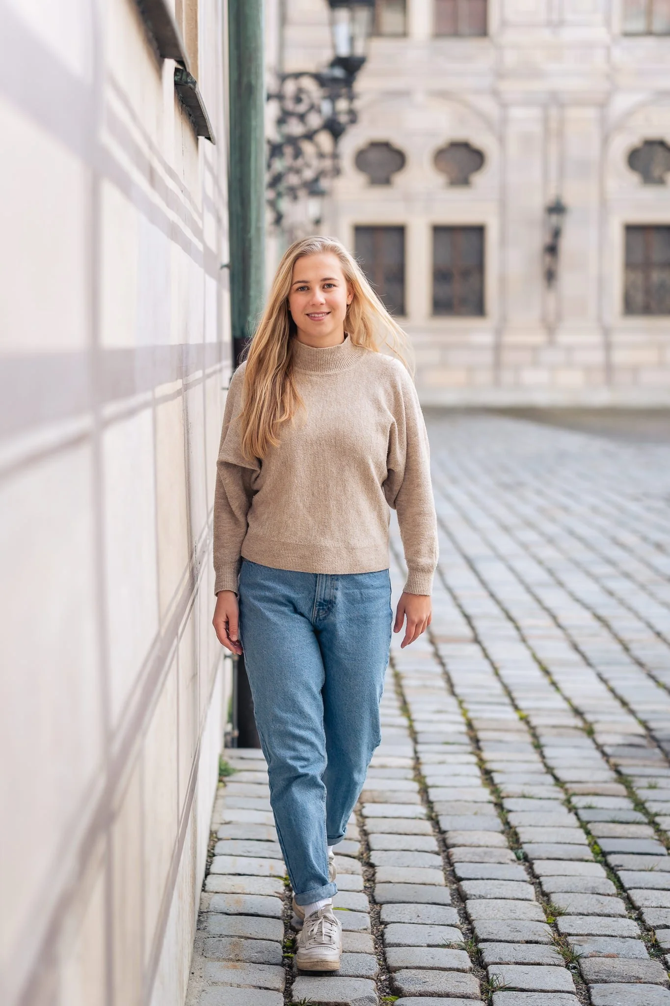 Junge Frau mit blonden Haaren, grauem Pullover, blauen Jeans und weißen Schuhen, geht die Kopfsteingasse entlang, hinter ihr ein historisches Gebäude