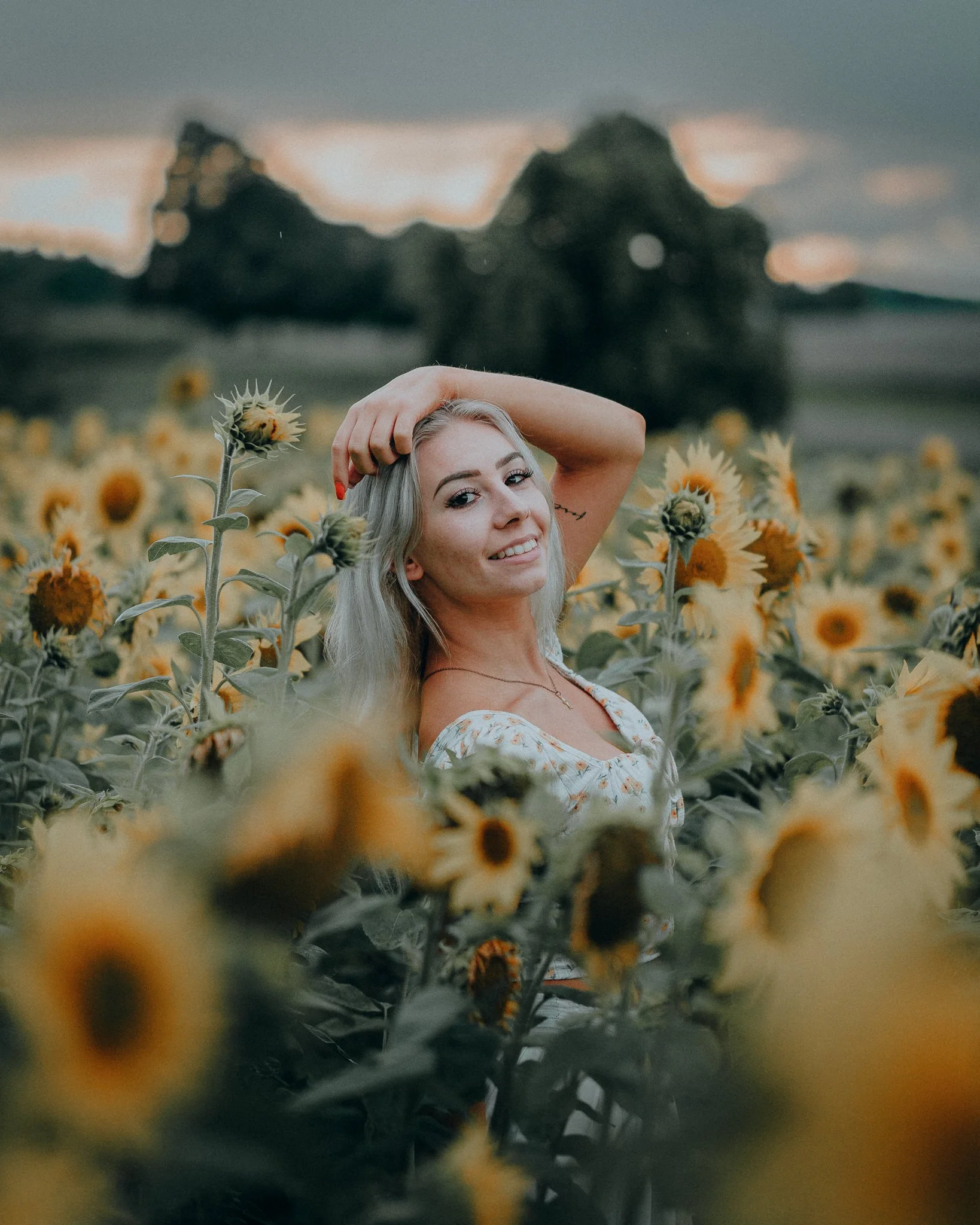 Eine junge Frau mit blonden Haaren posiert in einem Sonnenblumenfeld, während sie die Hand auf den Kopf legt und in die Kamera lächelt, mit weichem Licht im Hintergrund.
