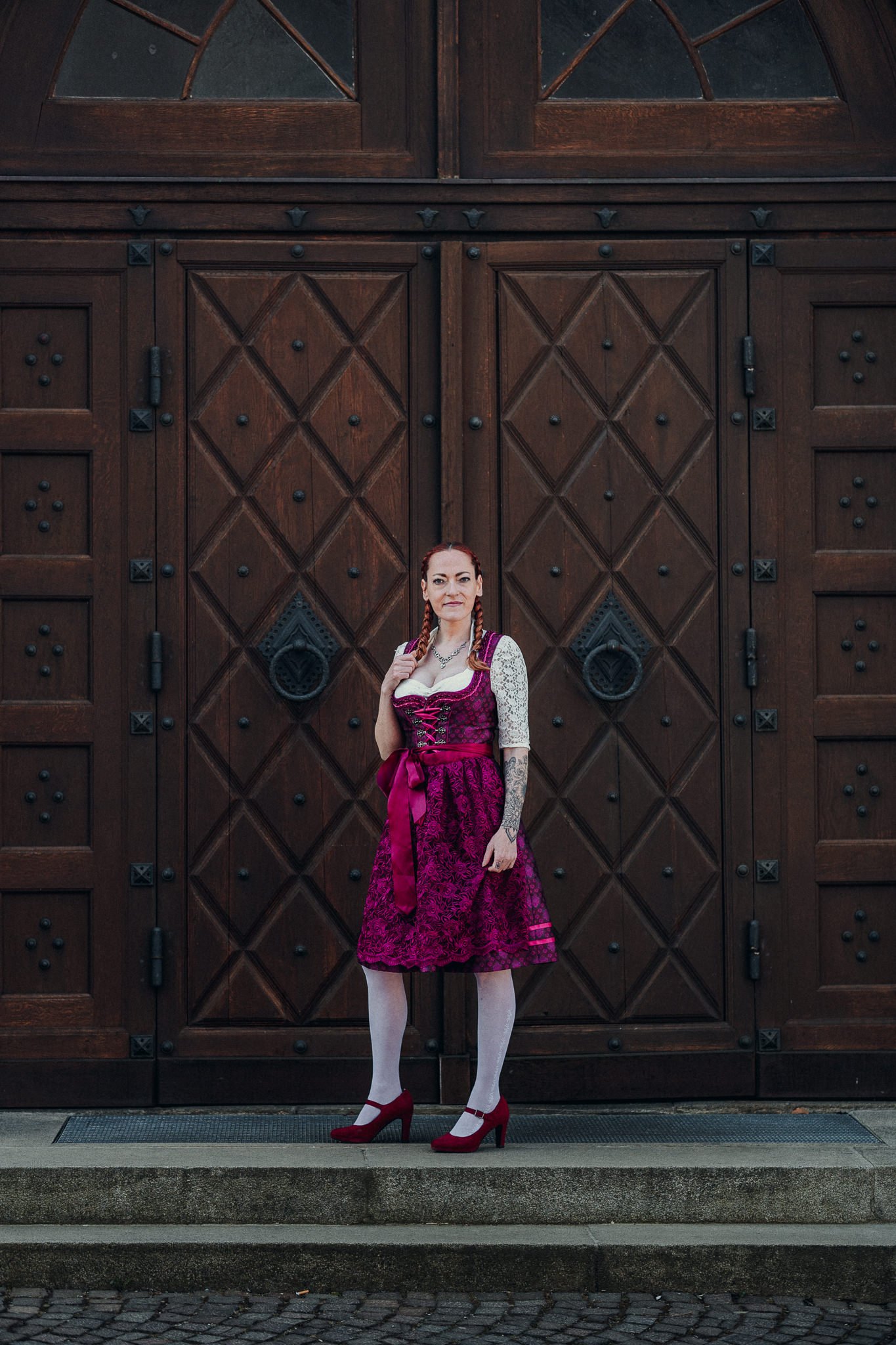 Frau in einem rosa-lila Dirndl mit weißen Strümpfen und roten Schuhen steht vor einer großen, dunklen Holztür.