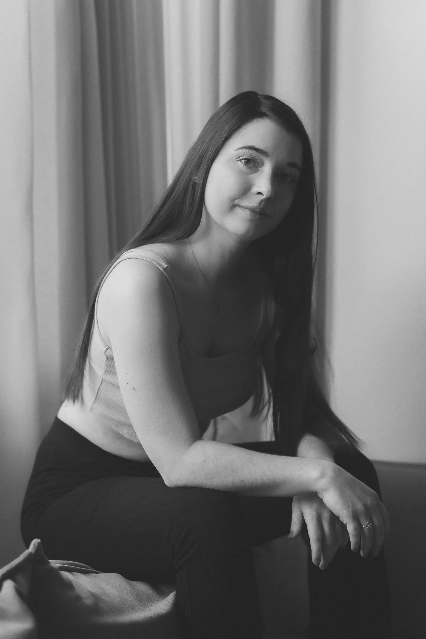 Schwarz-weiß-Foto einer jungen Frau mit langen, dunklen Haaren, sitzend vor einem Fenster mit Vorhängen, blickt lächelnd in die Kamera.