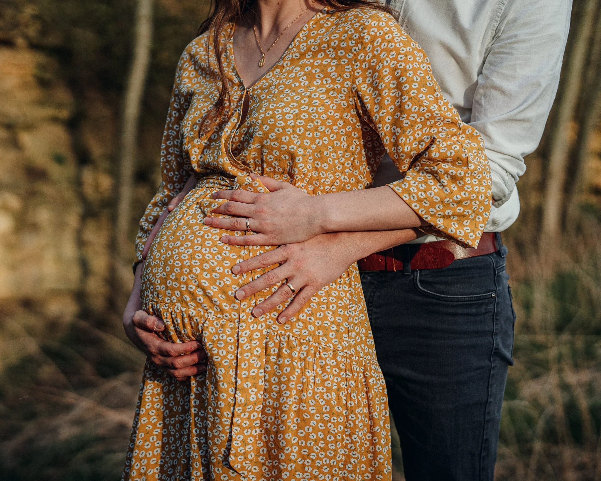 Ein schwangertes Paar umarmt sich im Freien, die Frau trägt ein gelbes Kleid mit weißen Mustern, die Hände liegen auf dem Babybauch.