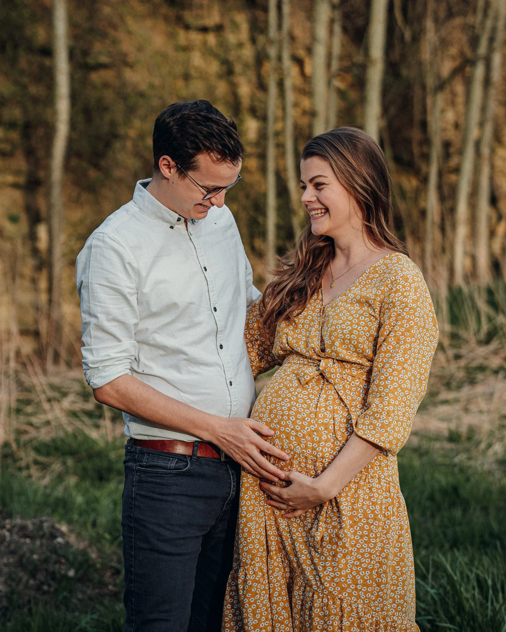 Ein glückliches, schwangres Paar steht im Freien, umgeben von Bäumen und Gras, während sie sich liebevoll an den Bauch fassen.