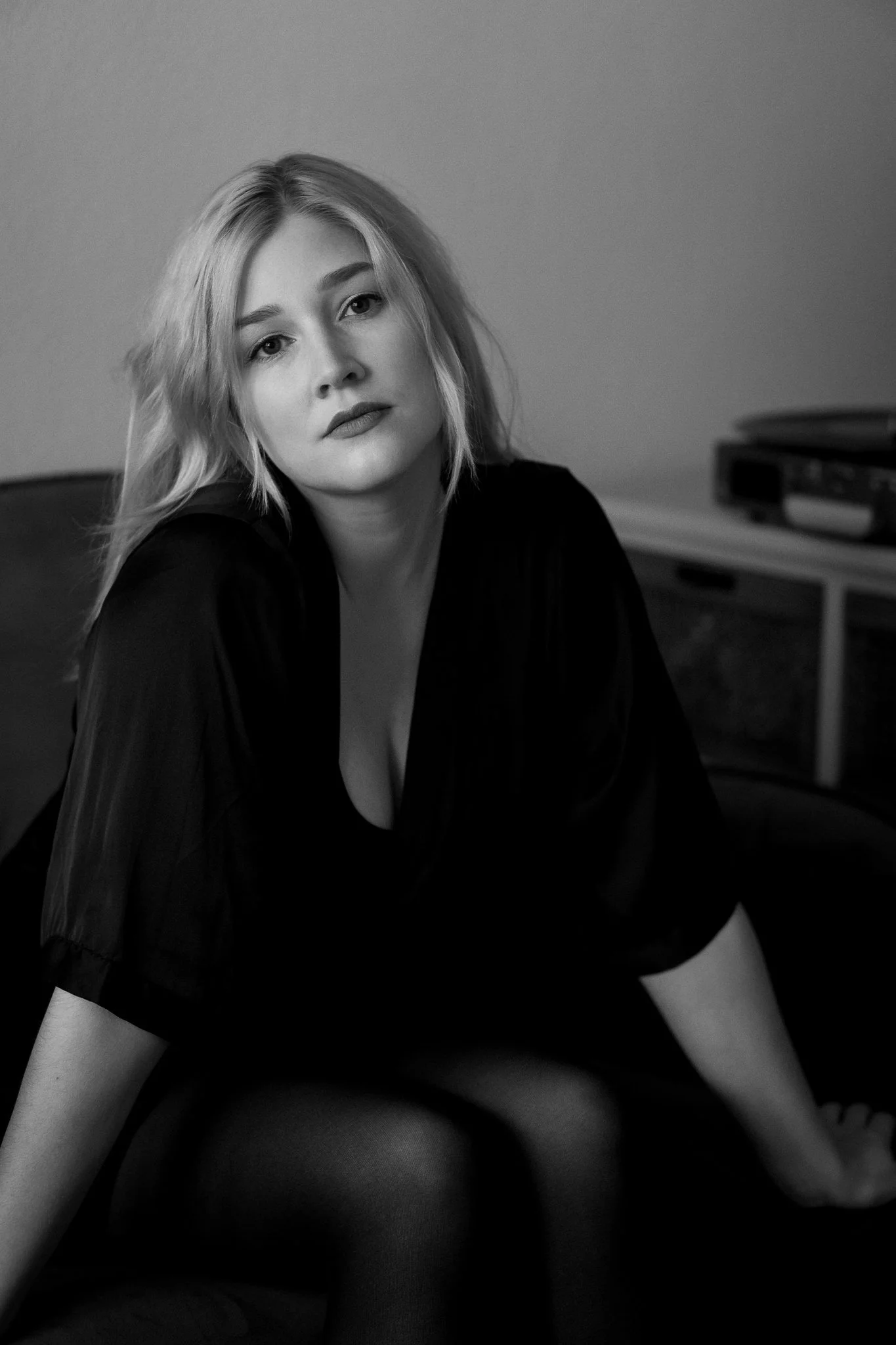 Schwarz-weiß Foto einer jungen Frau mit lockigem Haar, sitzend auf einem Sofa, trägt ein dunkles Kleid und schaut direkt in die Kamera.