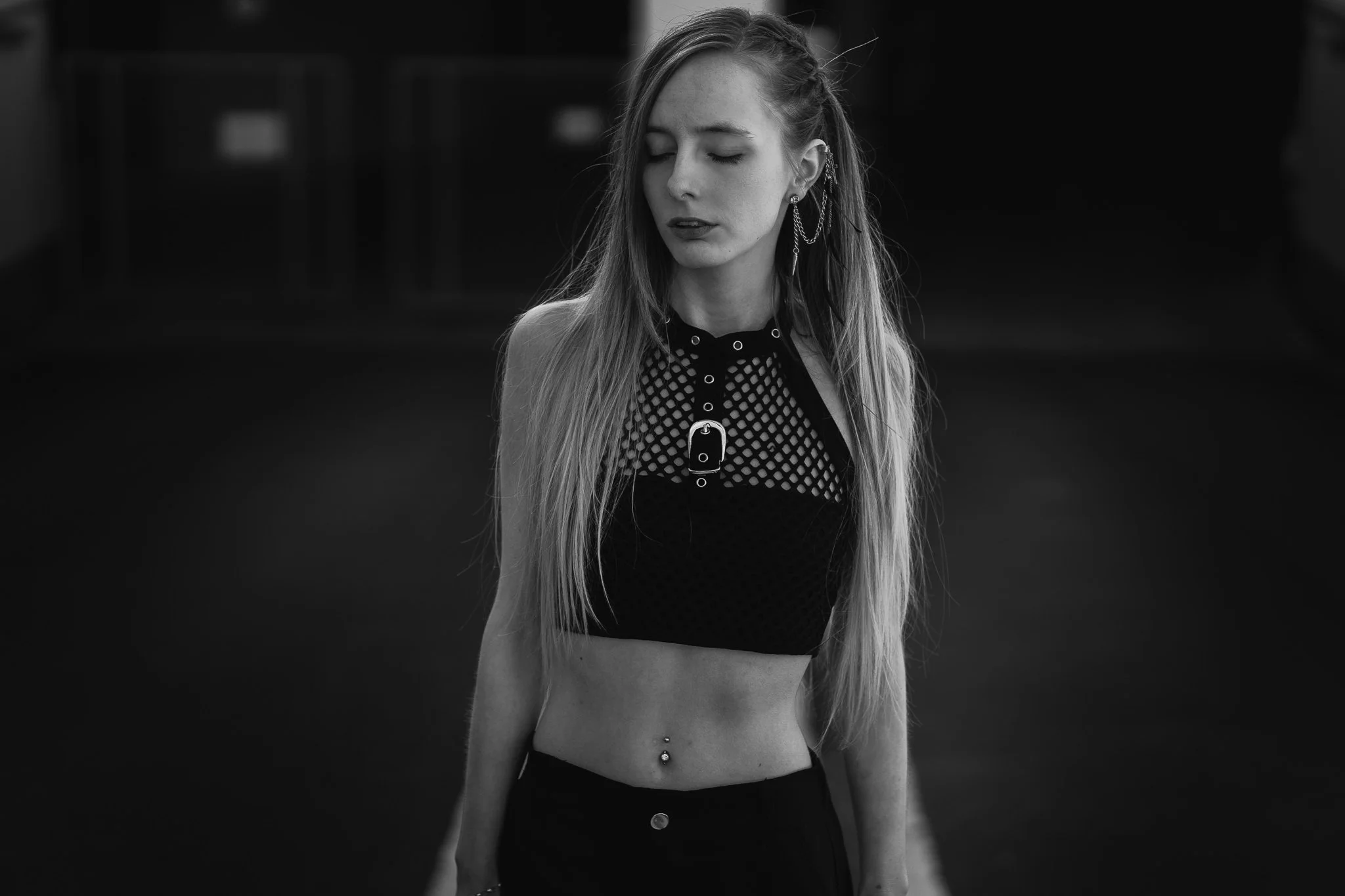 Schwarz-Weiß-Foto eines jungen Mädchens mit langen Haaren, das mit geschlossenen Augen nach links schaut, in einem dunklen Raum, trägt ein Netz-Top mit Leder-Details und einen Nabel-Piercing.