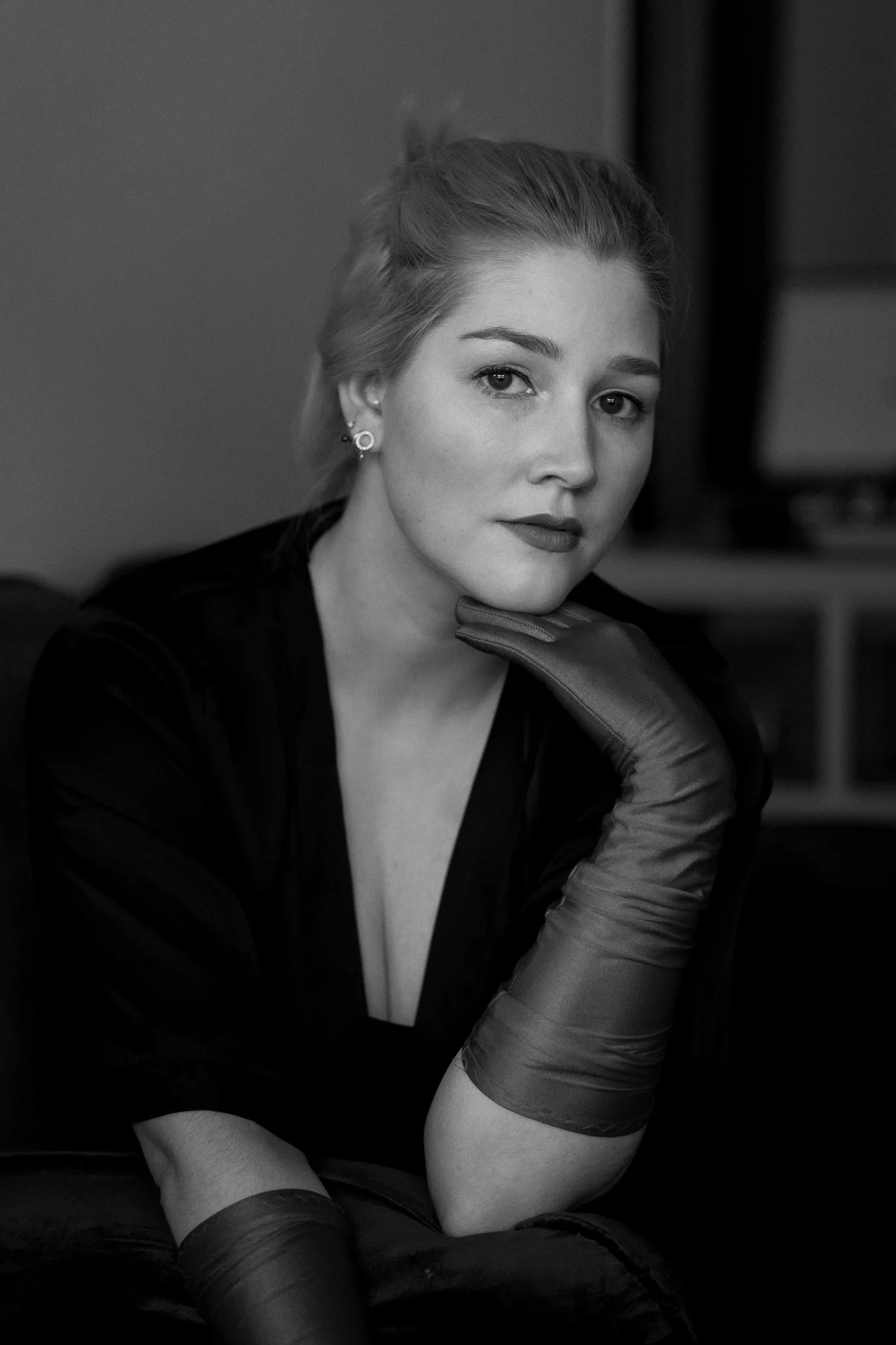 Schwarz-weiß Porträt einer jungen Frau mit blonder, glatter Frisur, dezenten Make-up, Ohrringen und schwarzen Kleidung, die mit einem Handballen leicht ans Kinn gestützt ist.