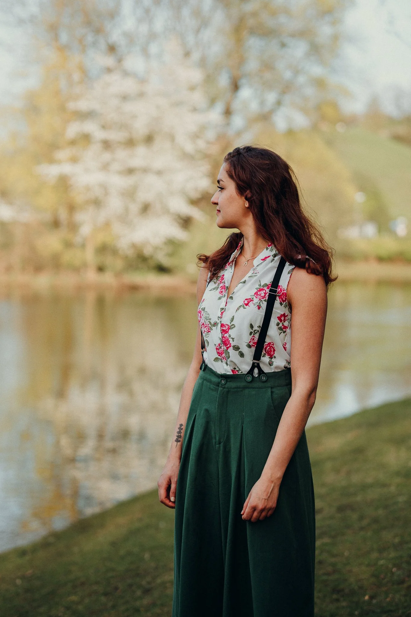 Eine Frau steht an einem Fluss mit Bäumen im Hintergrund, trägt eine weiße Bluse mit roten Rosenmustern und eine grüne Hose.