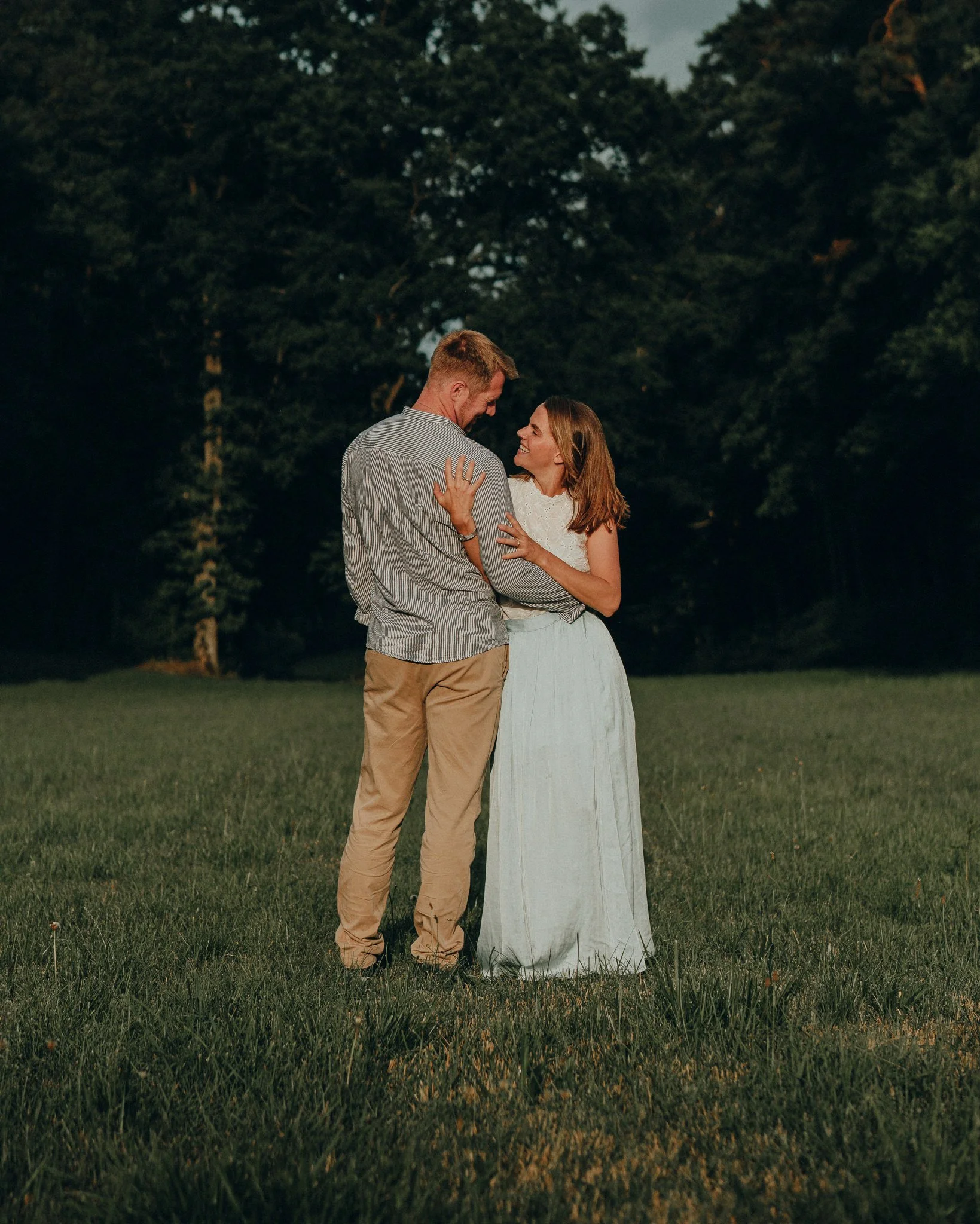 Ein Paar umarmt sich auf einer grünen Wiese vor einem dunklen Wald bei Sonnenuntergang.