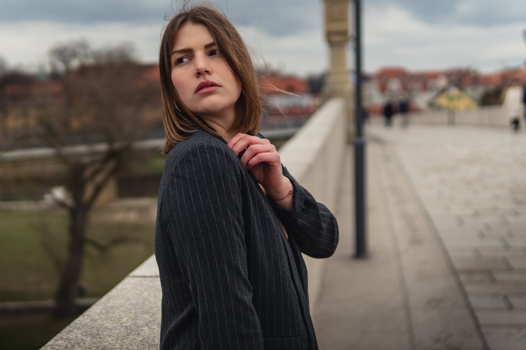 Junge Frau mit braunen Haaren und dunklem Anzug auf einer Brücke in einer europäischen Stadt, im Hintergrund Bäume und historische Gebäude, bewölkter Himmel.