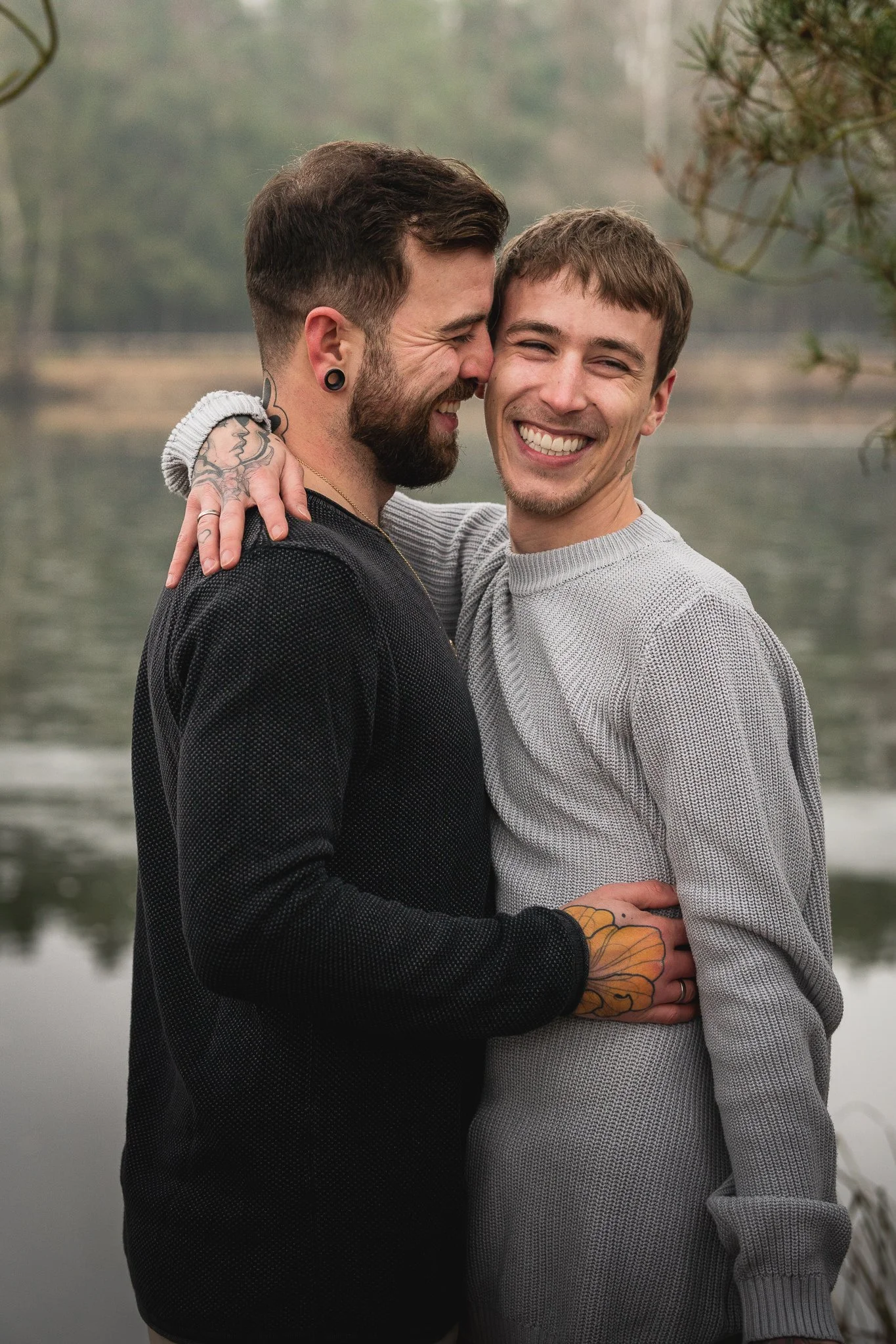 Zwei Männer umarmen sich lachend am See, im Hintergrund Bäume und Wasser.