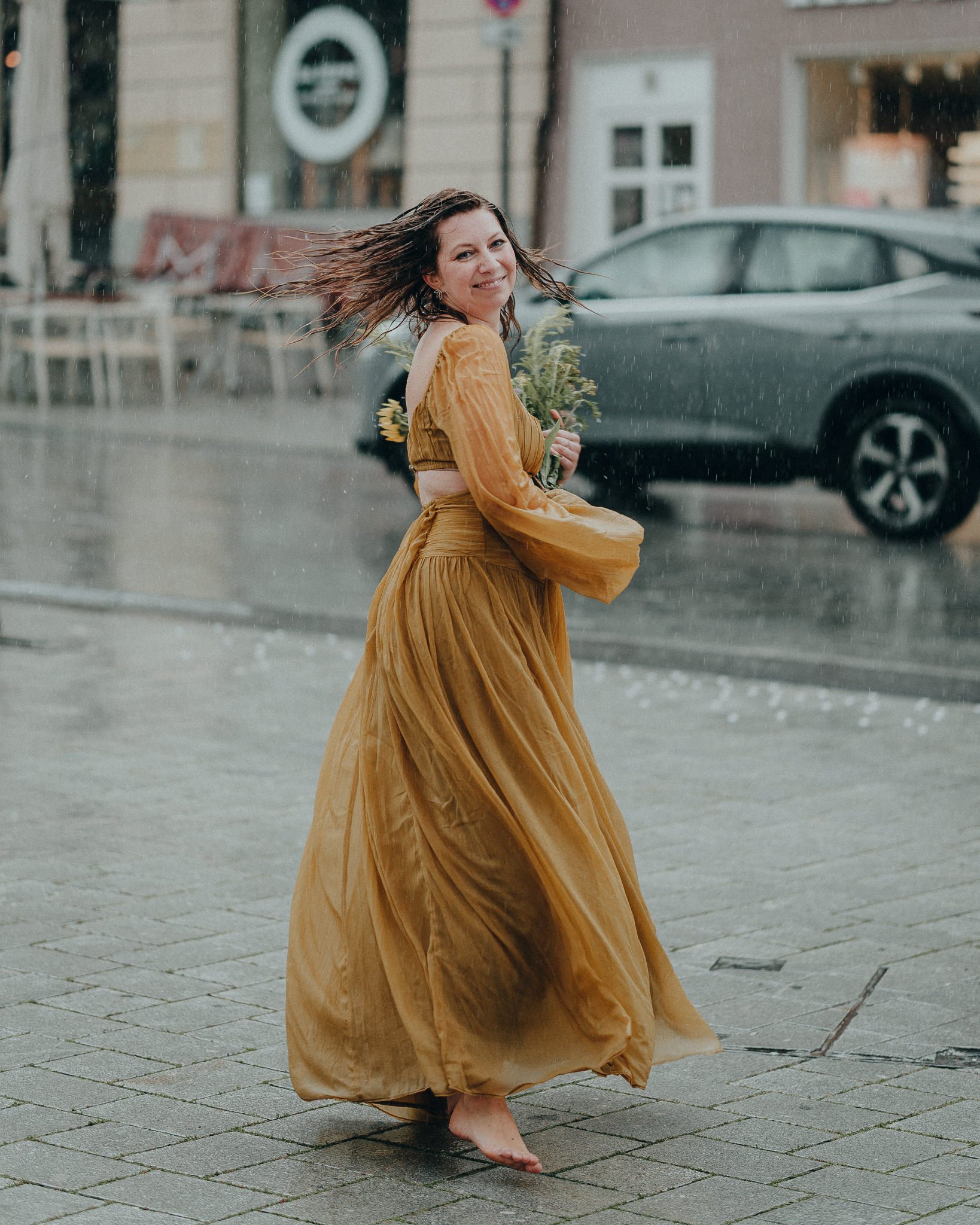 Eine lachende Frau in einem goldgelben Kleid läuft barfuß bei Regen auf einer Stadtstraße, hält einen Blumenstrauß und schaut zurück.