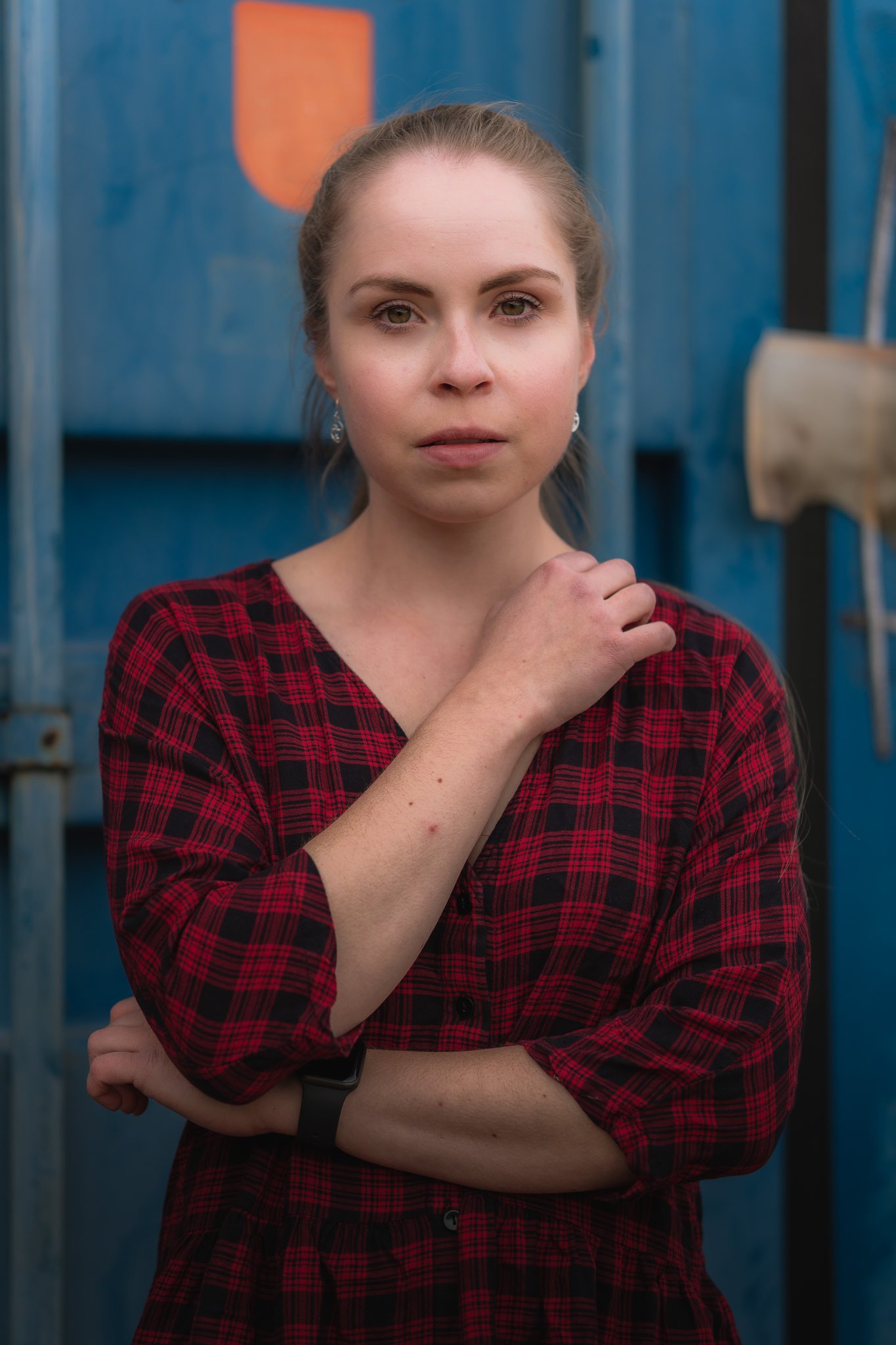 Junge Frau mit grauen Augen und braunem Haar in Zopf, trägt rote Karierte Bluse, steht vor blauer Metallstruktur, wirkt nachdenklich.