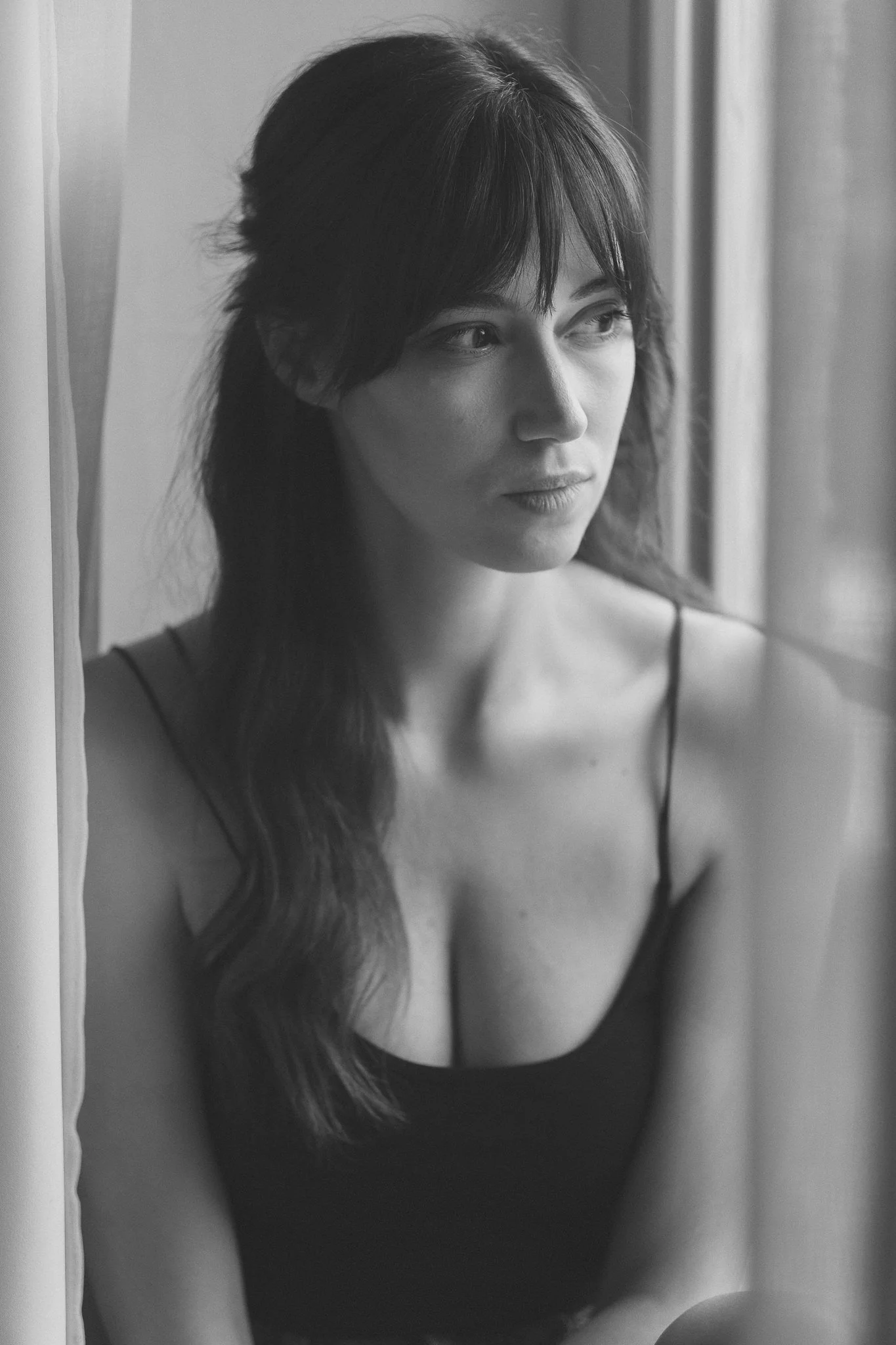Schwarz-Weiß-Fotografie einer jungen Frau mit mittellangen, leicht gewellten Haaren, die in die Ferne blickt, während sie neben einem Fenster sitzt.