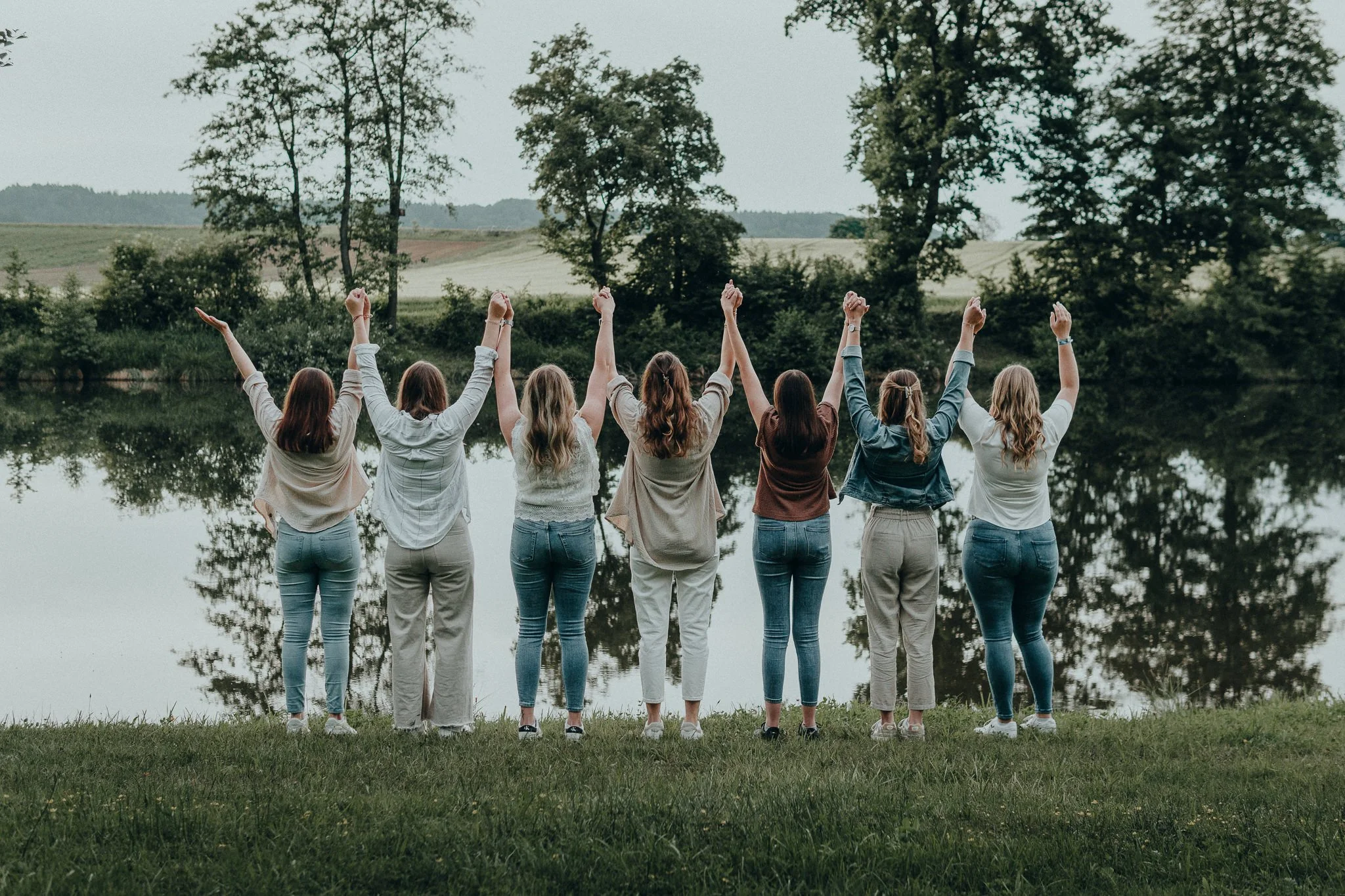 Sieben Frauen stehen am Ufer eines Sees, ihre Rücken sind sichtbar, und sie heben die Arme in die Luft, mit Blick auf die Natur und die Wasseroberfläche.