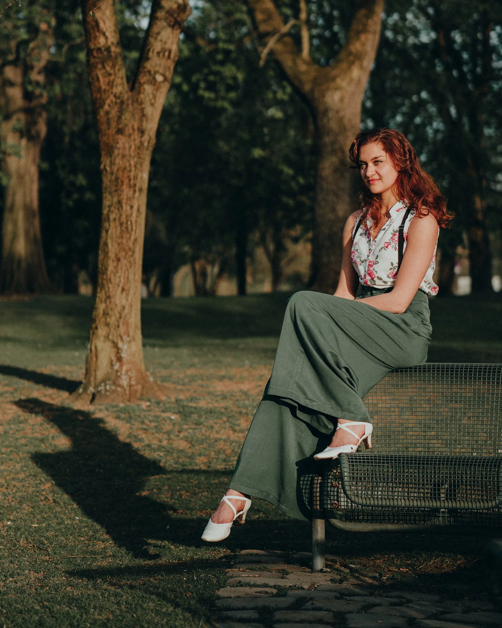 Eine junge Frau mit roten Haaren sitzt auf einer Parkbank im Grünen, trägt ein floral bedrucktes Top, eine lange grüne Hose und weiße Schuhe, hinter ihr sind große Bäume im Park sichtbar.