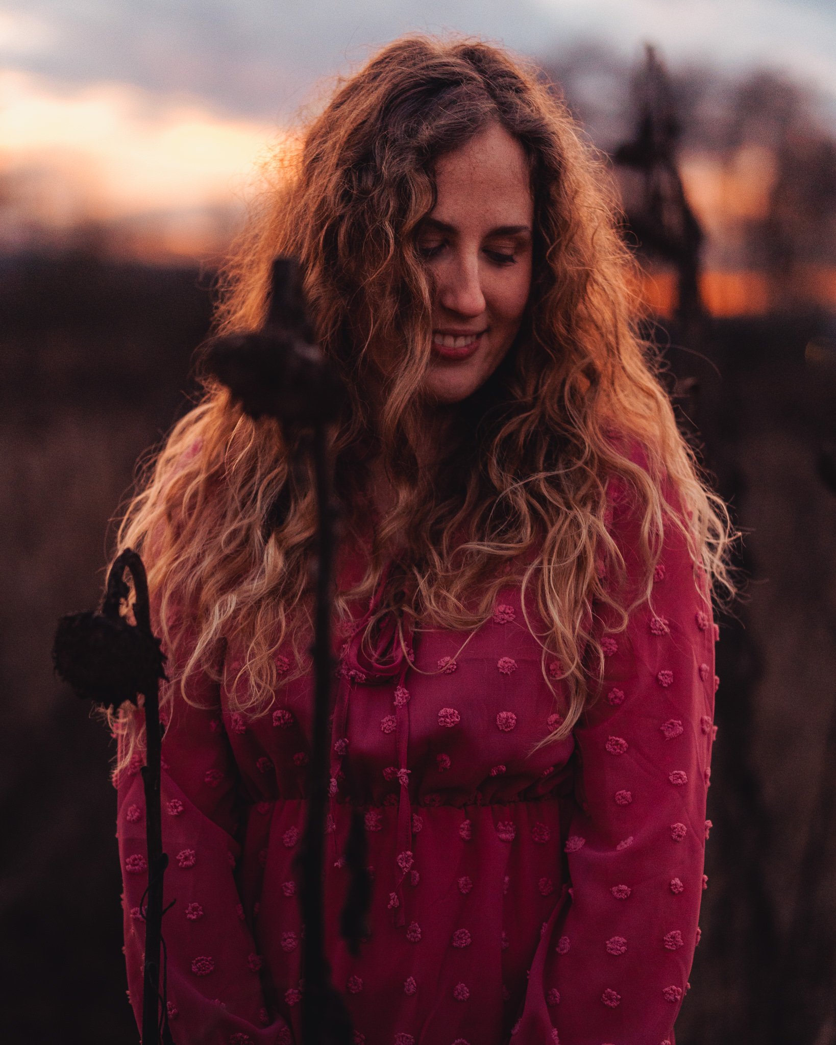 Eine junge Frau mit lockigem, rotem Haar trägt ein rotes Kleid mit Mustern, während sie im Freien bei Sonnenuntergang steht.