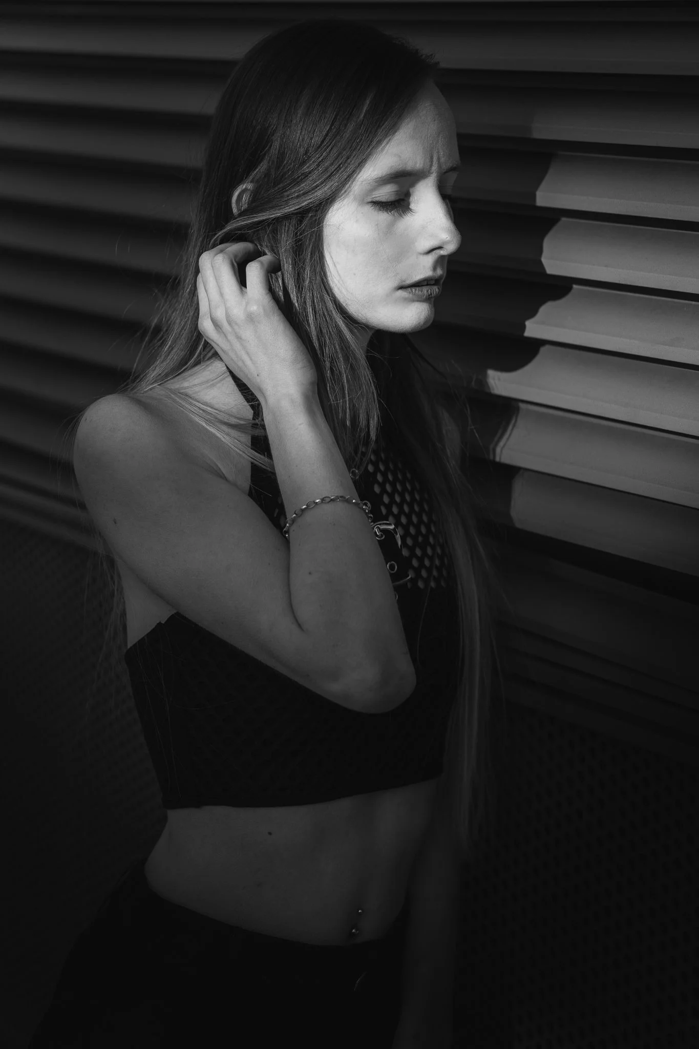 Schwarz-weiße Aufnahme einer jungen Frau mit langen Haaren, die die Augen geschlossen hat, während sie eine Hand an ihr Ohr legt. Sie steht vor einer Wand mit waagerechten Linien.