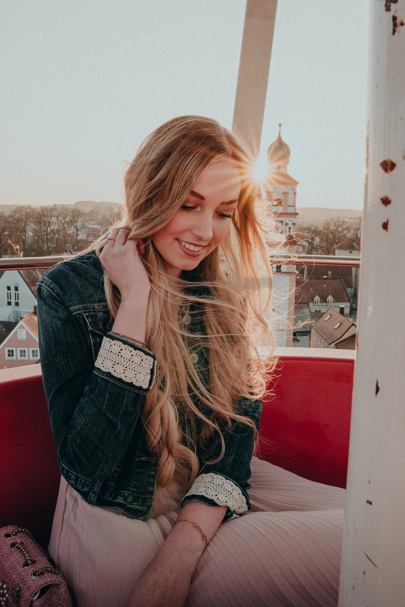 Junge Frau mit langen blonden Haaren, lachend, sitzt auf einer Dachterrasse bei Sonnenuntergang. Im Hintergrund sind Häuser und eine Kirche mit goldenem Dom sichtbar.
