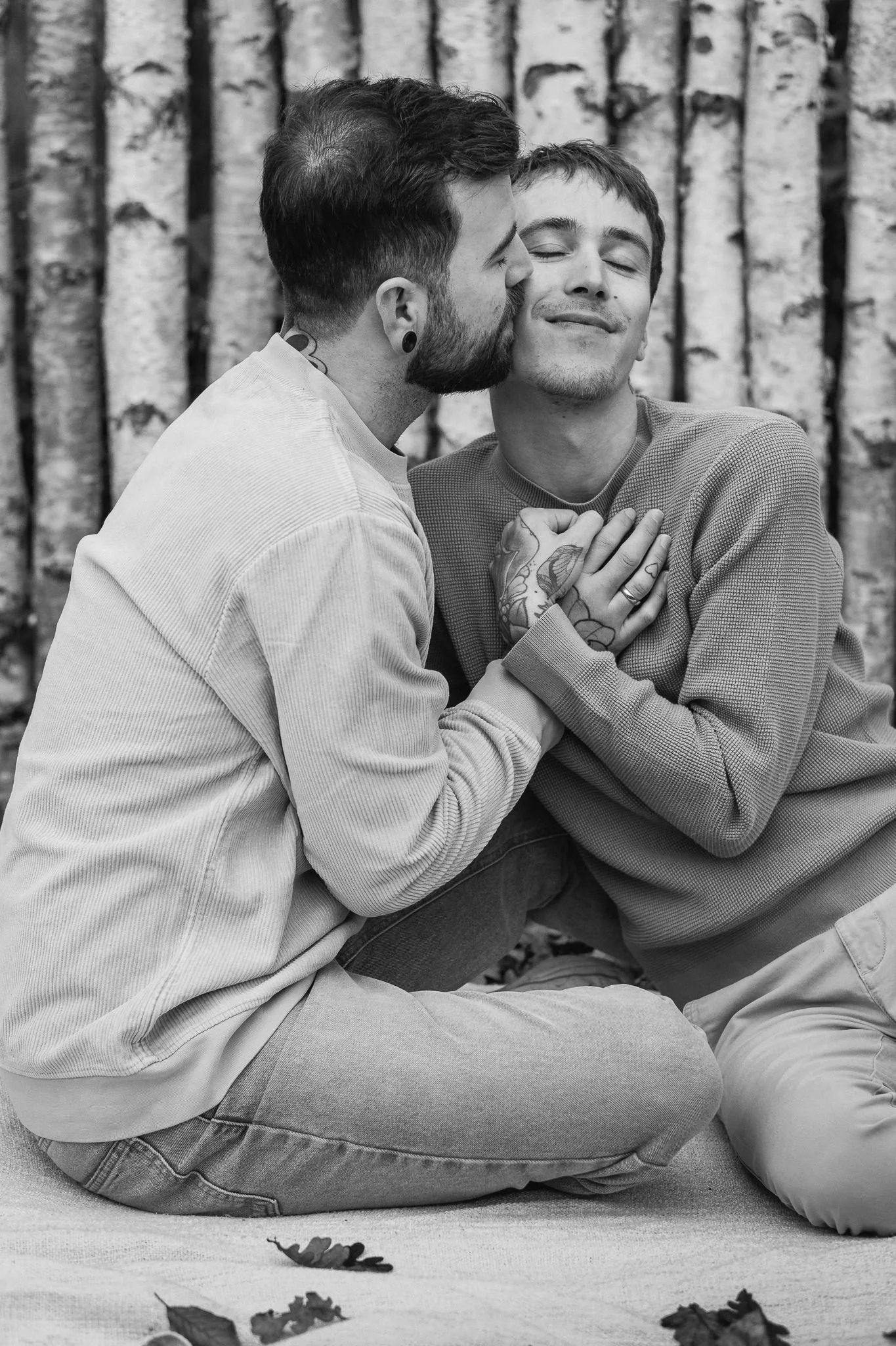 Zwei Männer sitzen auf einer Decke im Wald, einer küsst den anderen auf die Wange, beide lächeln glücklich, im Hintergrund Bäume.