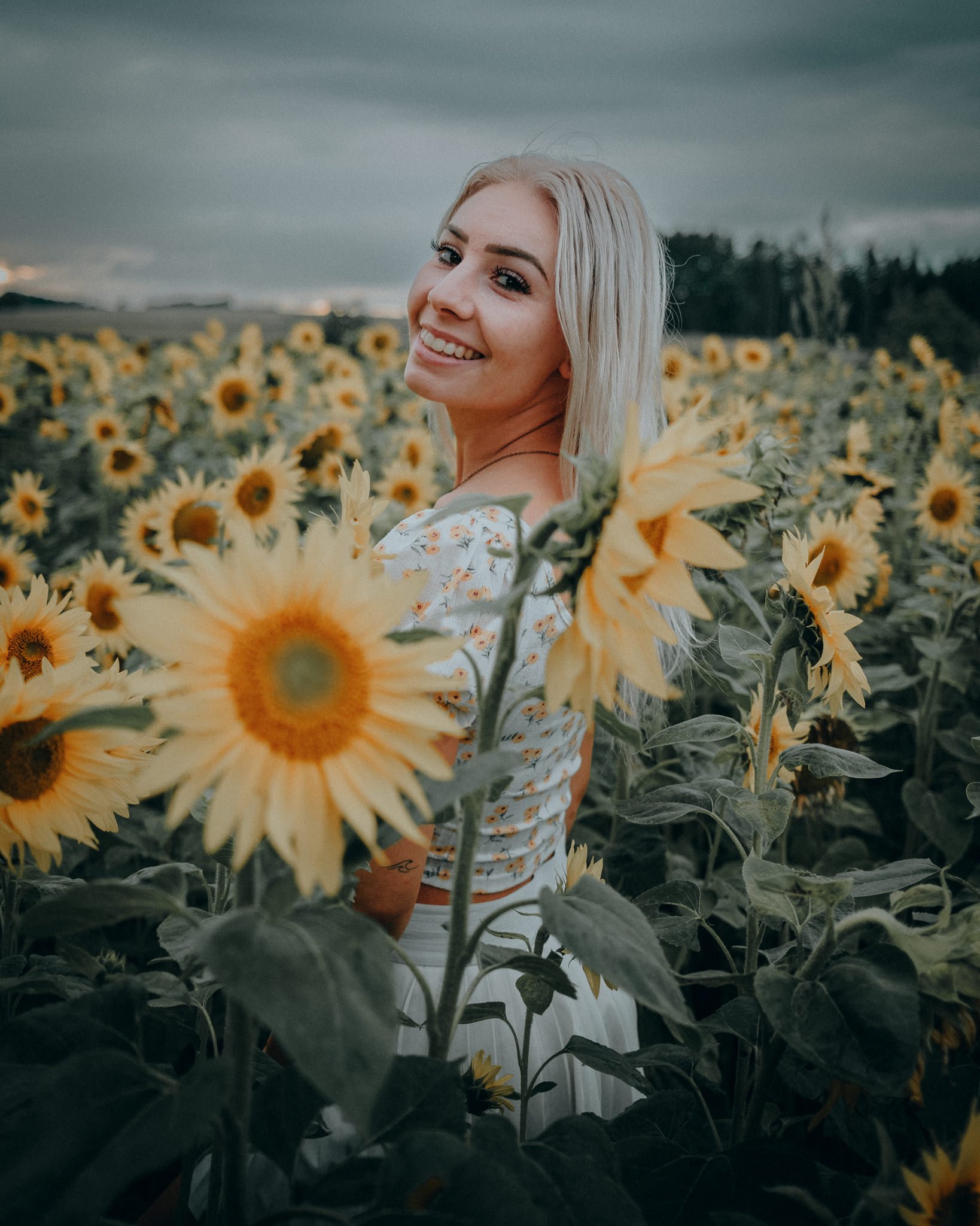 Eine lachende blonde Frau steht in einem Sonnenblumenfeld unter bewölktem Himmel.