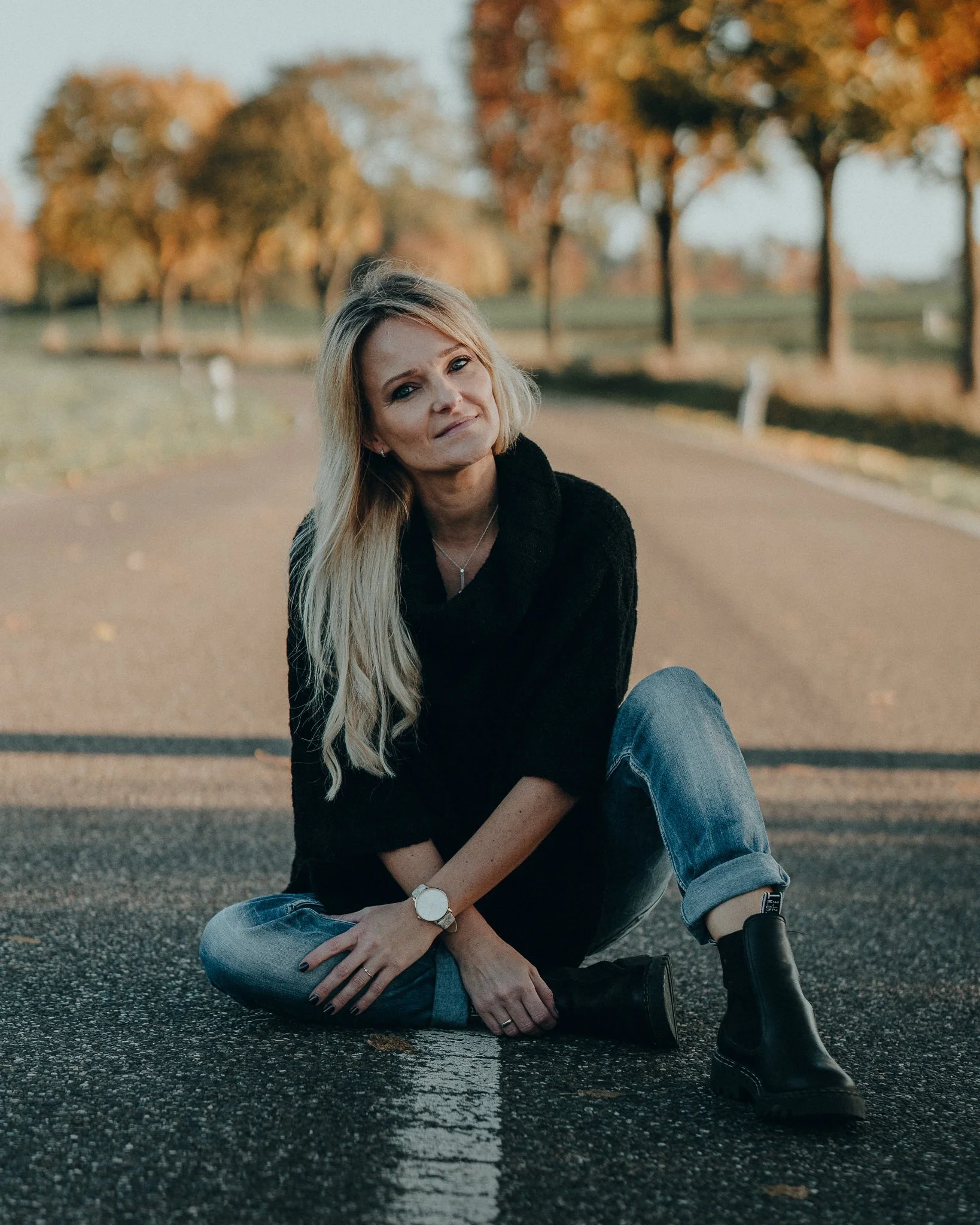 Eine blonde Frau sitzt auf einer Straße im Freien, umgeben von Bäumen mit herbstlichen Blättern, trägt schwarze Kleidung und Jeans, und schaut nach oben.