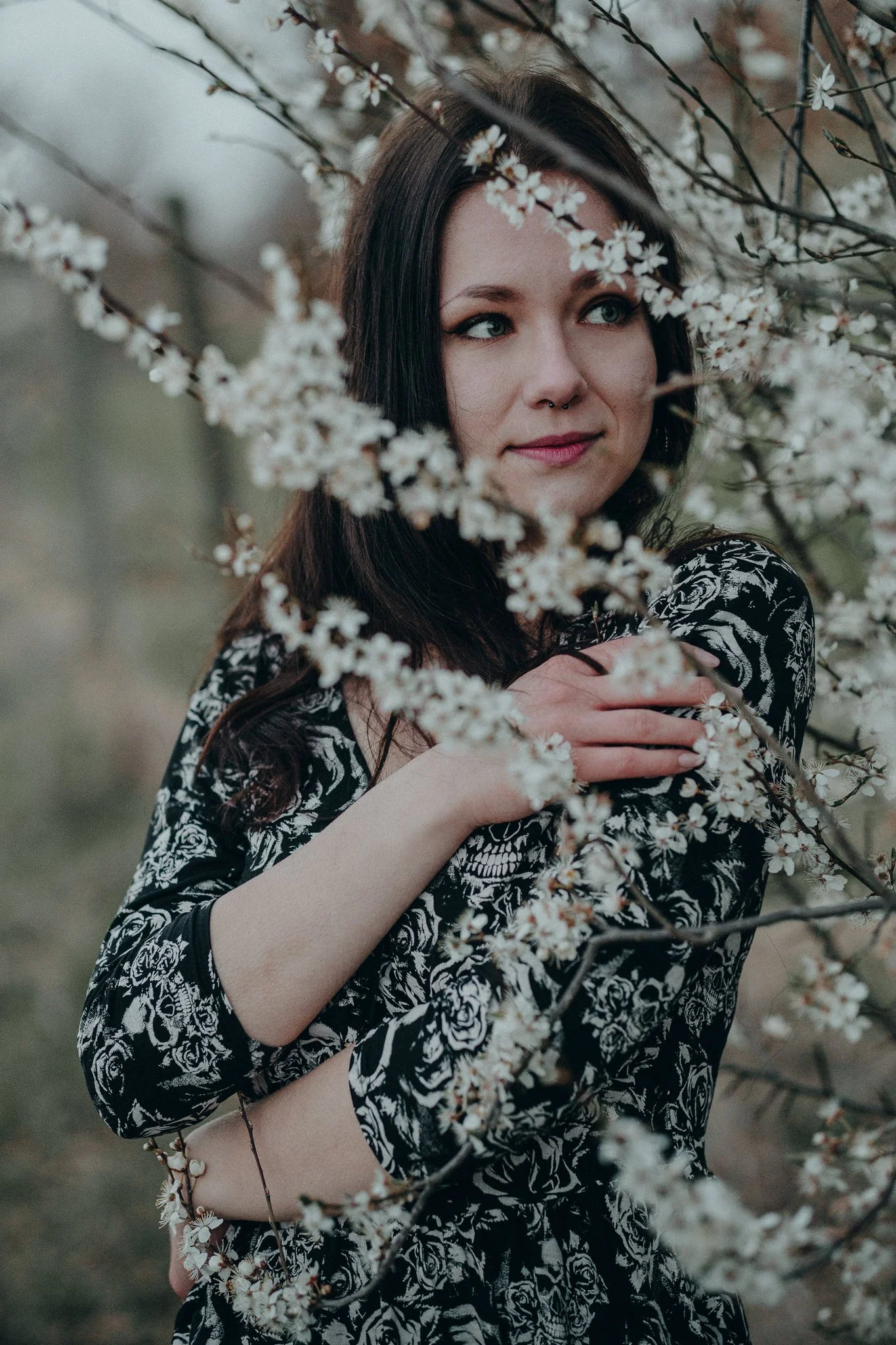 Junge Frau mit schwarzen Haaren und Nasenpiercing, umgeben von blühenden Zweigen, trägt ein schwarzes Kleid mit weißen Rosenmustern.