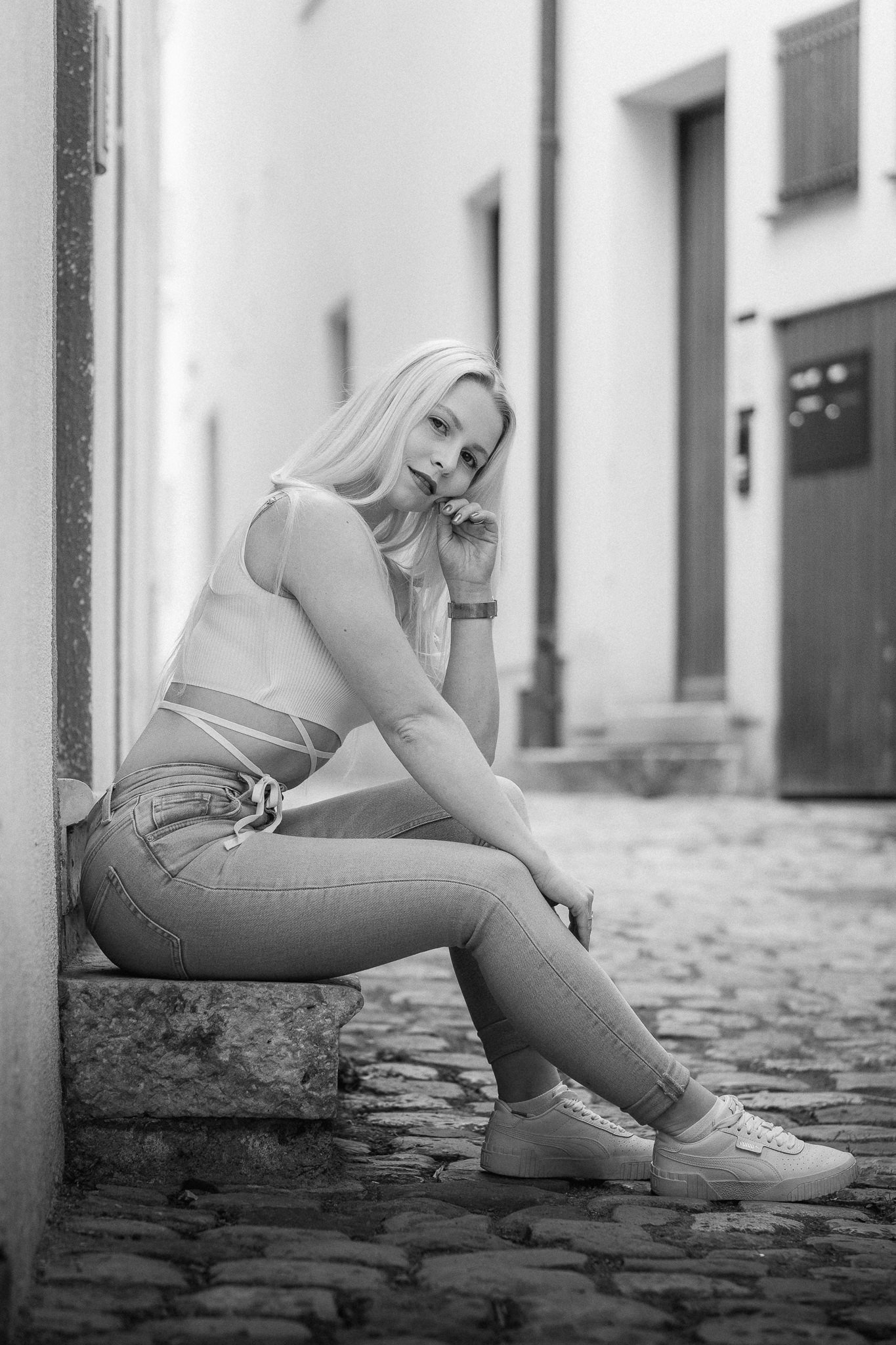Junge Frau sitzt auf einem Stein an einer Kopfsteinpflasterstraße, trägt Jeans und Turnschuhe, lächelnd, mit hand vor dem Gesicht, in Schwarz-Weiß-Fotografie.