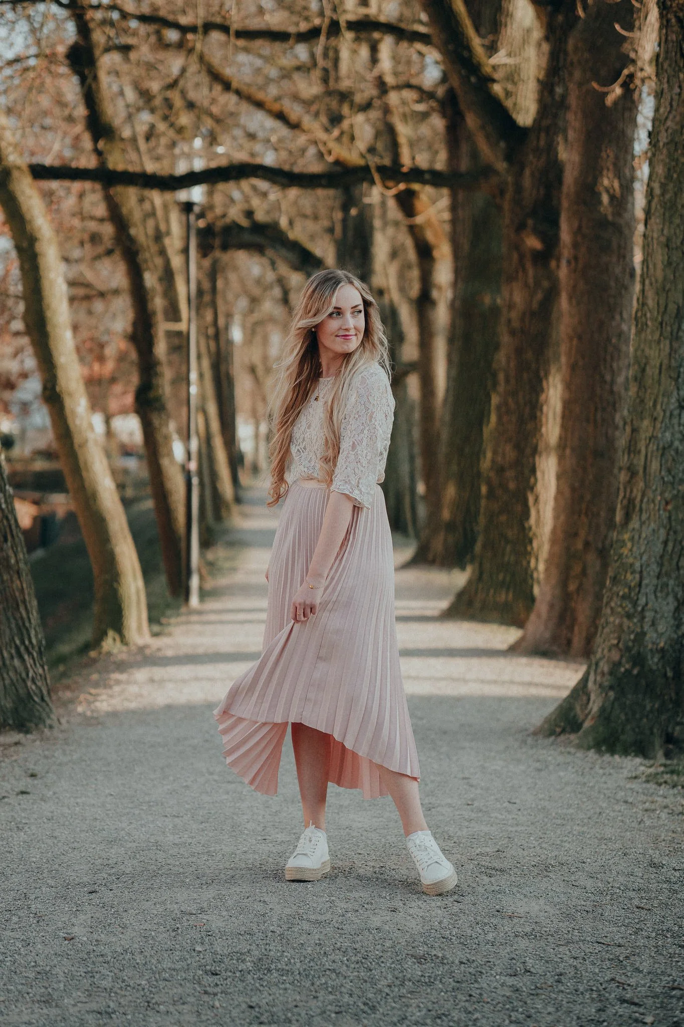 Eine junge Frau in einem weißen Spitzenblusen und einer hellrosa, plissierten Midirock steht auf einem Gehweg unter Bäumen, die im Herbst ihre Blätter verloren haben. Sie trägt weiße Sneakers und schaut leicht lächelnd zur Seite.