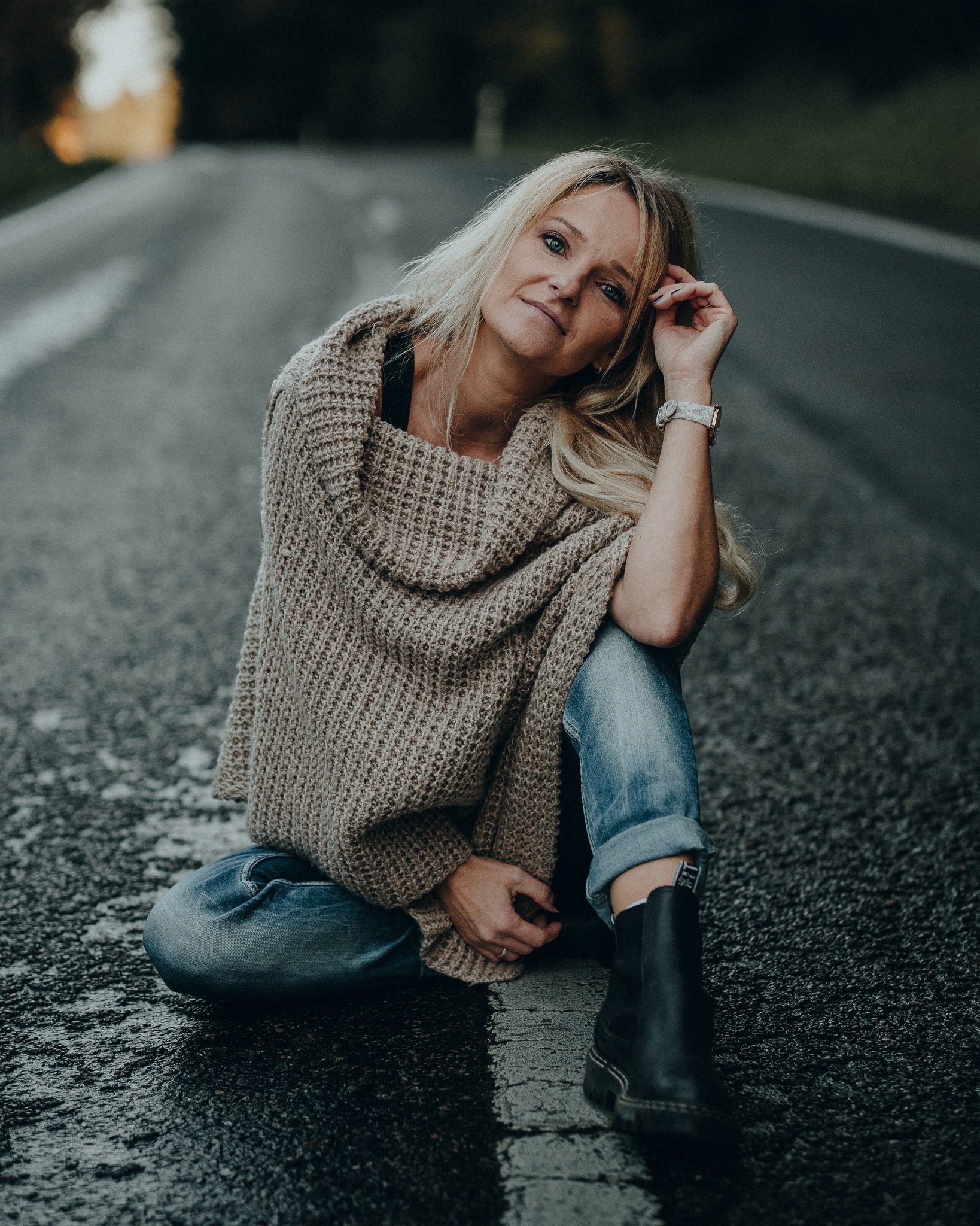 Eine Frau sitzt auf einer nassen Straße, trägt einen groben, beigen Pullover, Jeans und schwarze Stiefel, mit einem nachdenklichen Gesichtsausdruck.