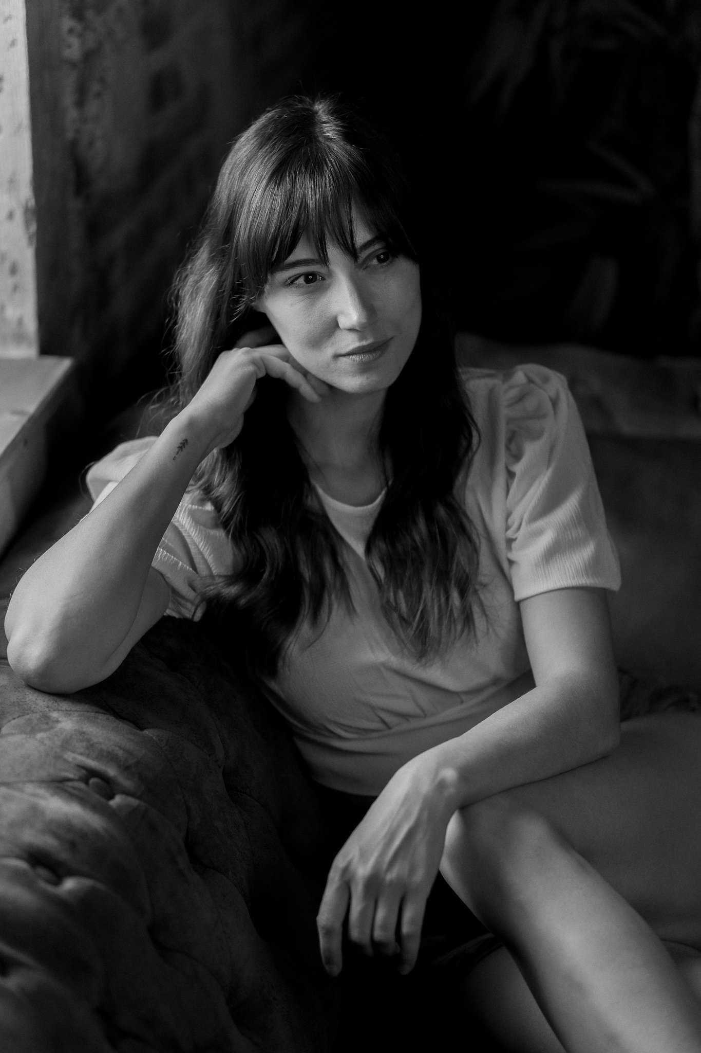 Schwarz-weiß-Fotografie einer jungen Frau, die entspannt auf einer Couch sitzt, mit langem, dunklem Haar und einem nachdenklichen Ausdruck.