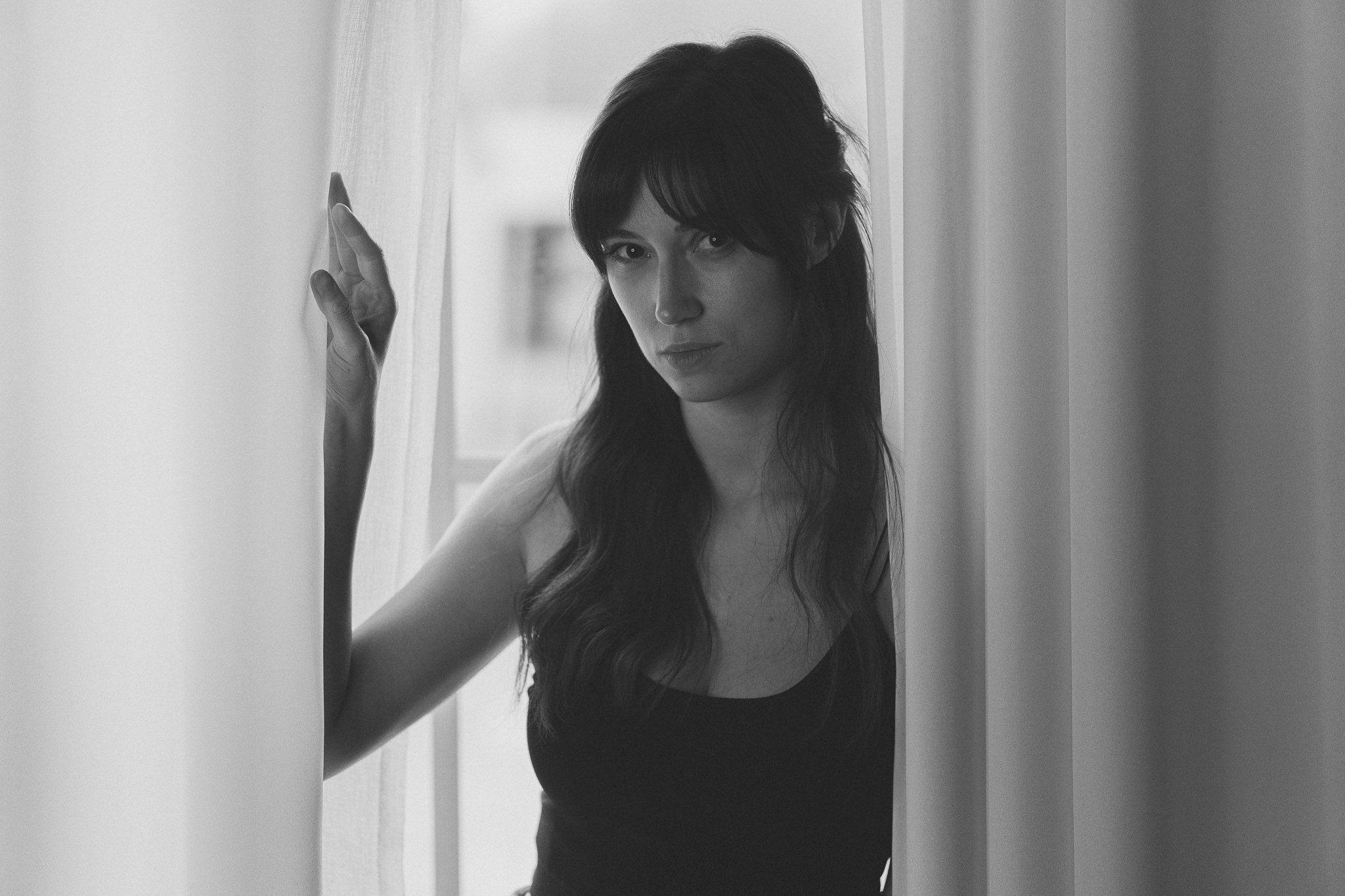 Schwarz-Weiß-Foto einer jungen Frau mit dunklen, gewellten Haaren, die durch einen Türvorhang blickt, mit ernstem Gesichtsausdruck.