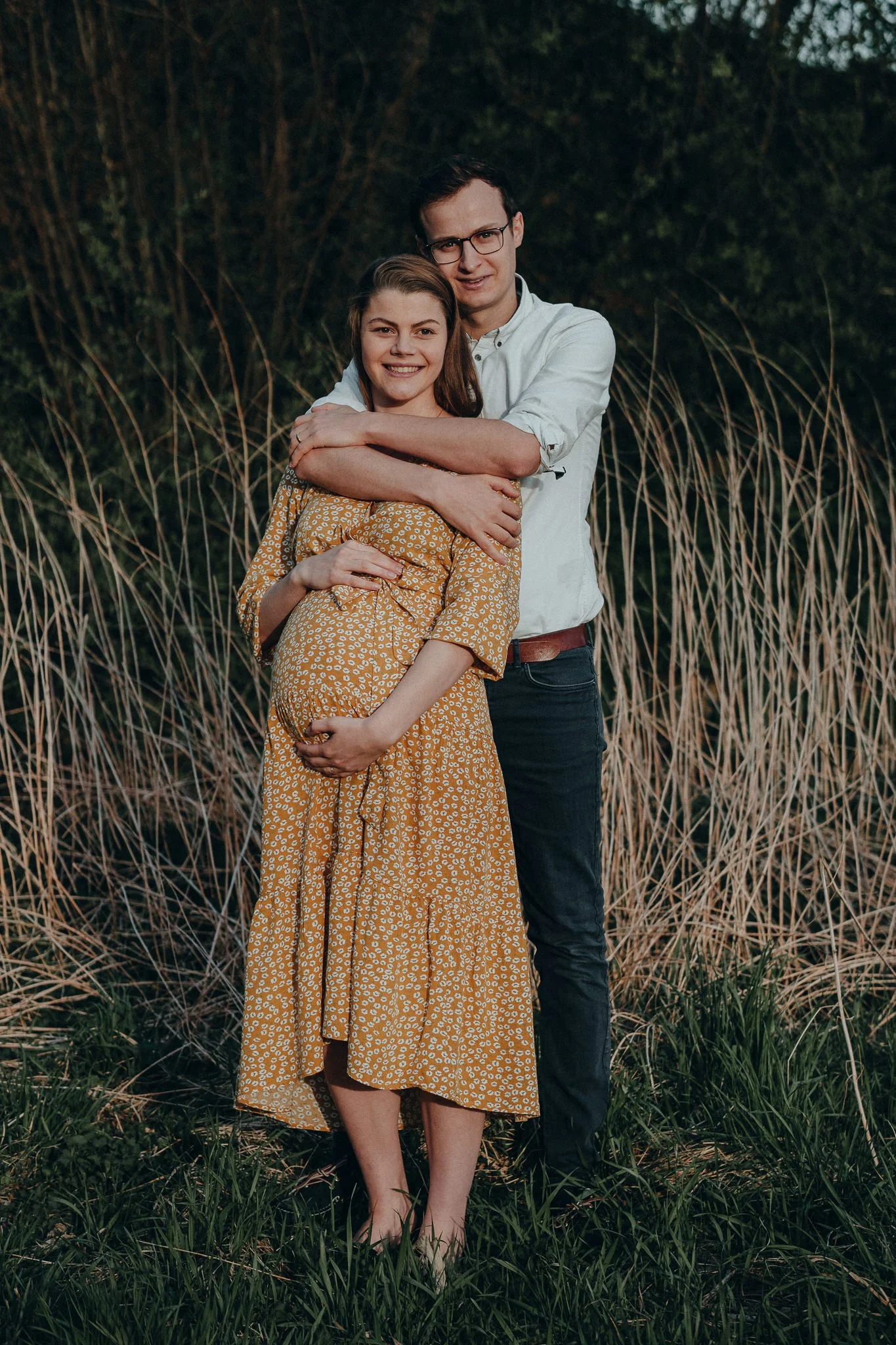 Ein Paar steht im Freien in einem Feld mit hohen Gräsern, die Frau trägt ein gelbes Kleid mit weißen Mustern, der Mann trägt ein weißes Hemd und dunkle Hose, beide lächeln und schauen in die Kamera.