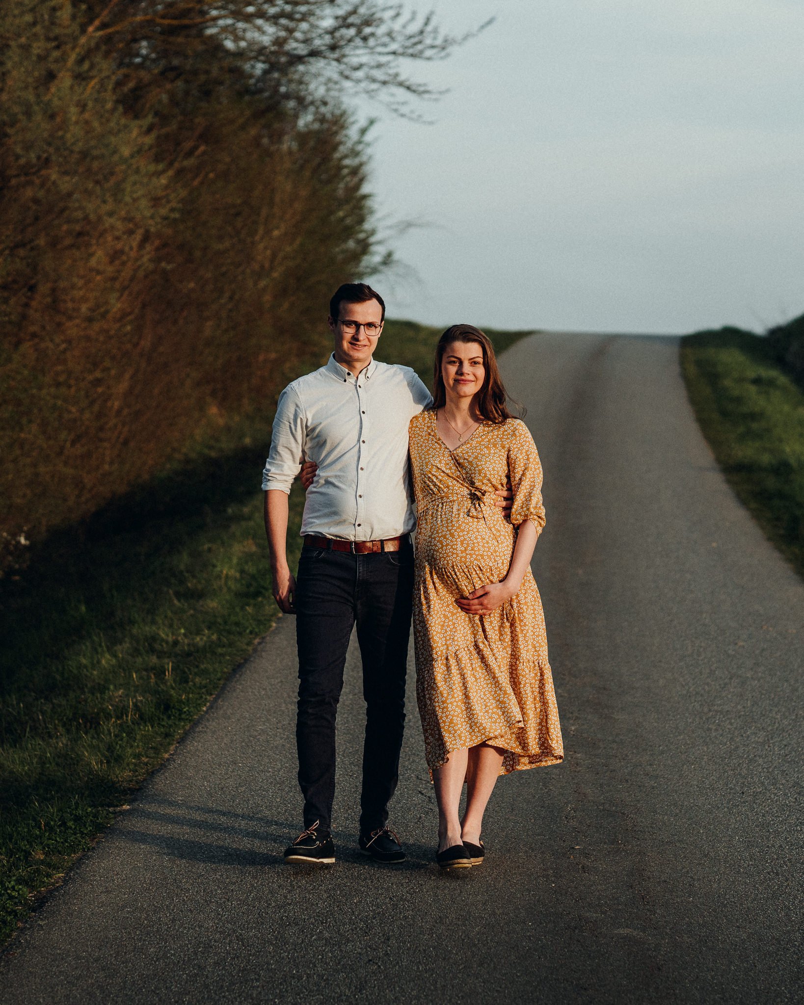 Ein Paar, ein Mann und eine schwangere Frau, gehen Hand in Hand auf einer ländlichen Straße bei Sonnenuntergang.