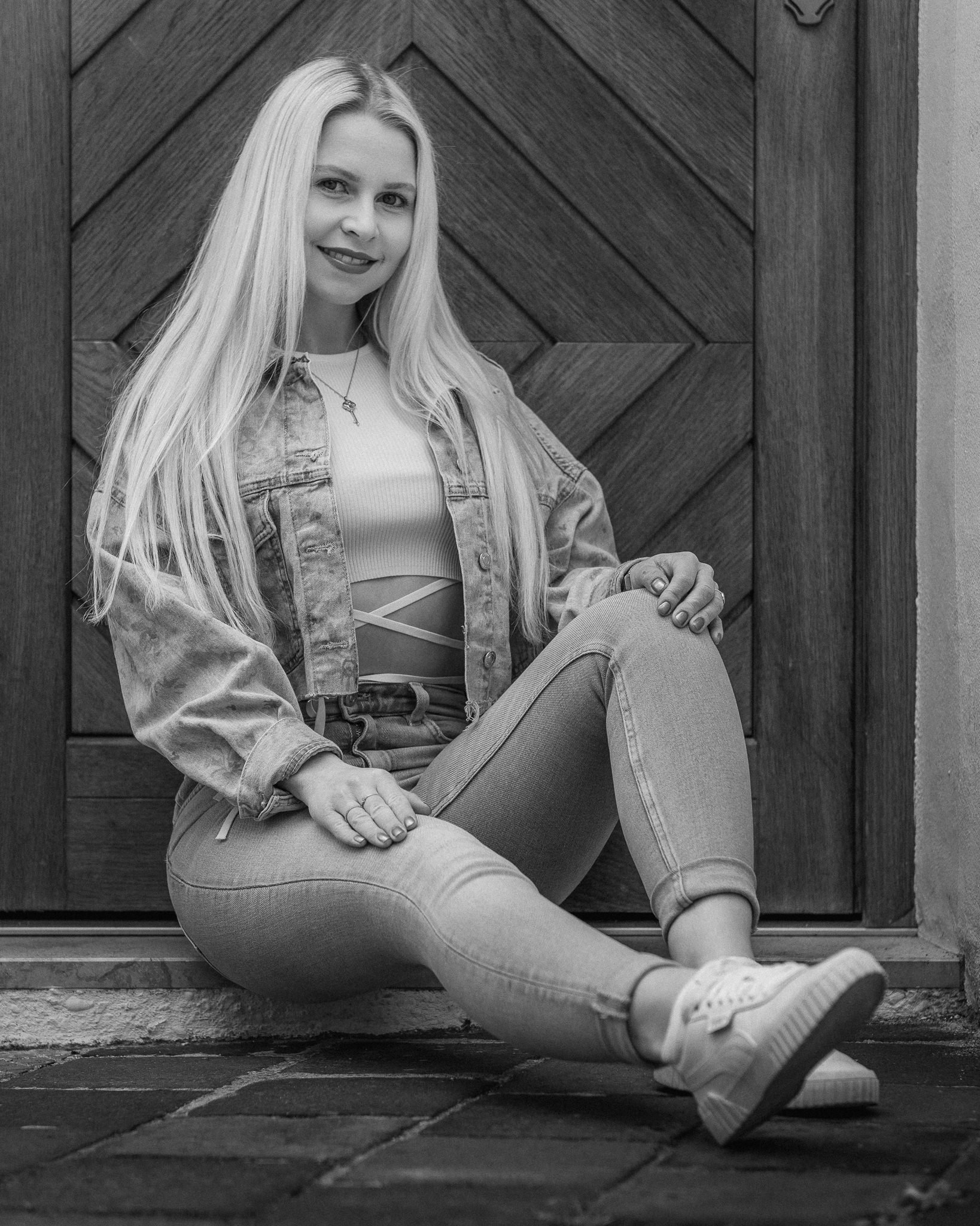 Junge Frau sitzt auf dem Boden vor einer Holztür, trägt Jeans, Turnschuhe und eine Jacke, lächelt und schaut in die Kamera.