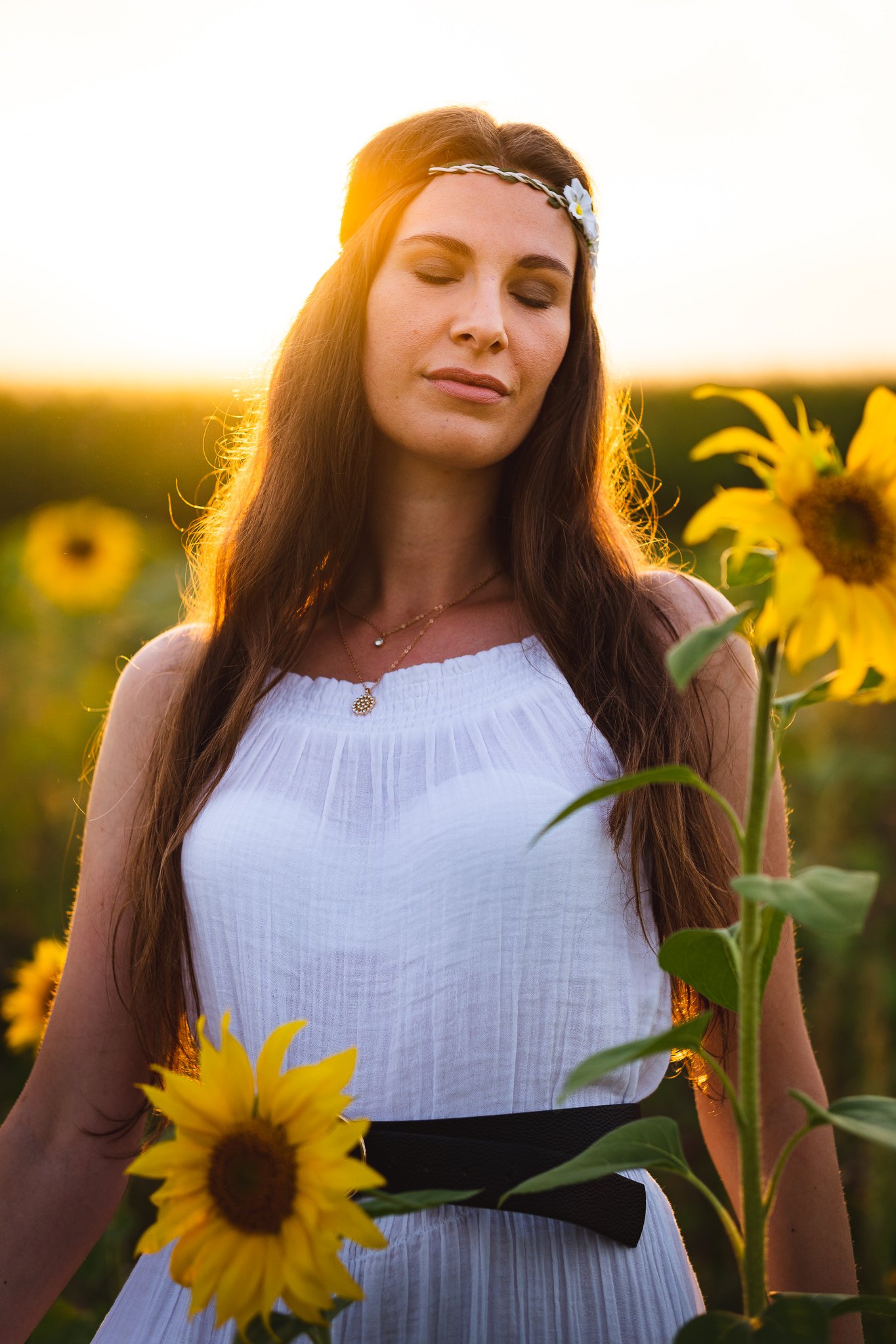 Eine junge Frau mit langen braunen Haaren, weißem Kleid und Blume im Haar steht in einem Sonnenblumenfeld bei Sonnenuntergang.