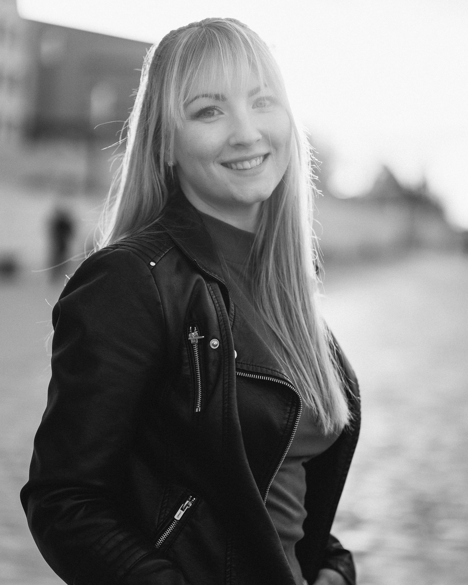 Schwarz-Weiß-Foto einer lachenden jungen Frau mit langem Haar, die eine Lederjacke trägt, im Freien bei Sonnenuntergang.