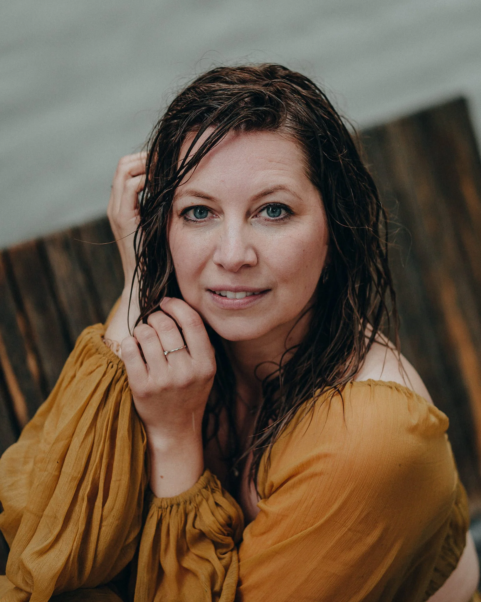 Nahaufnahmen einer Frau mit nassem, dunklem Haar, und blauen Augen, die ein gelbliches Oberteil trägt, sitzt auf einer Holzbank. Sie schaut direkt in die Kamera.