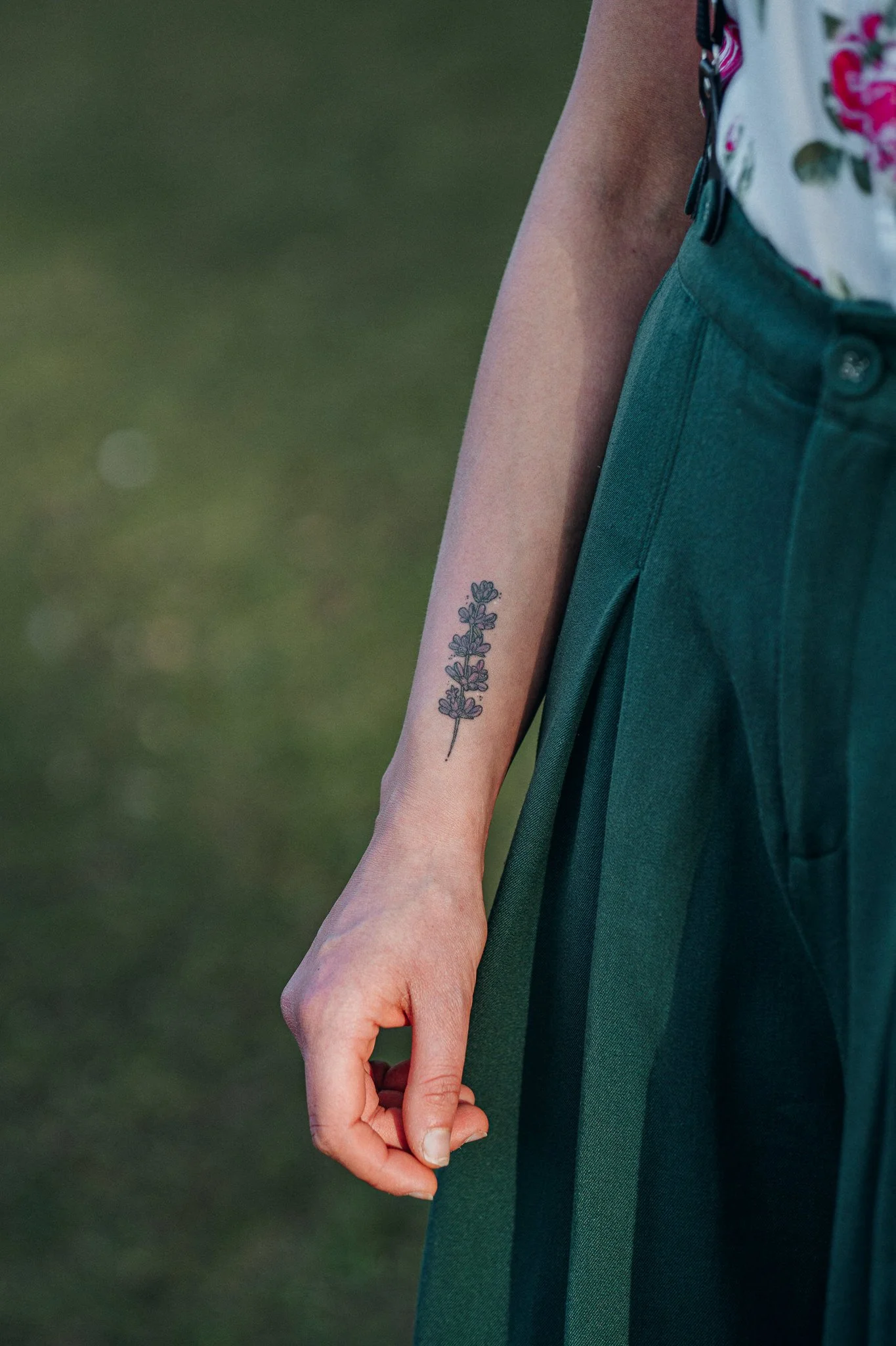 Nahaufnahme von einem Arm mit Tätowierung einer Lavendelpflanze, Kleidung mit grünen Stoff und schwarzem Streifen, im Hintergrund unscharfer grüner Rasen.