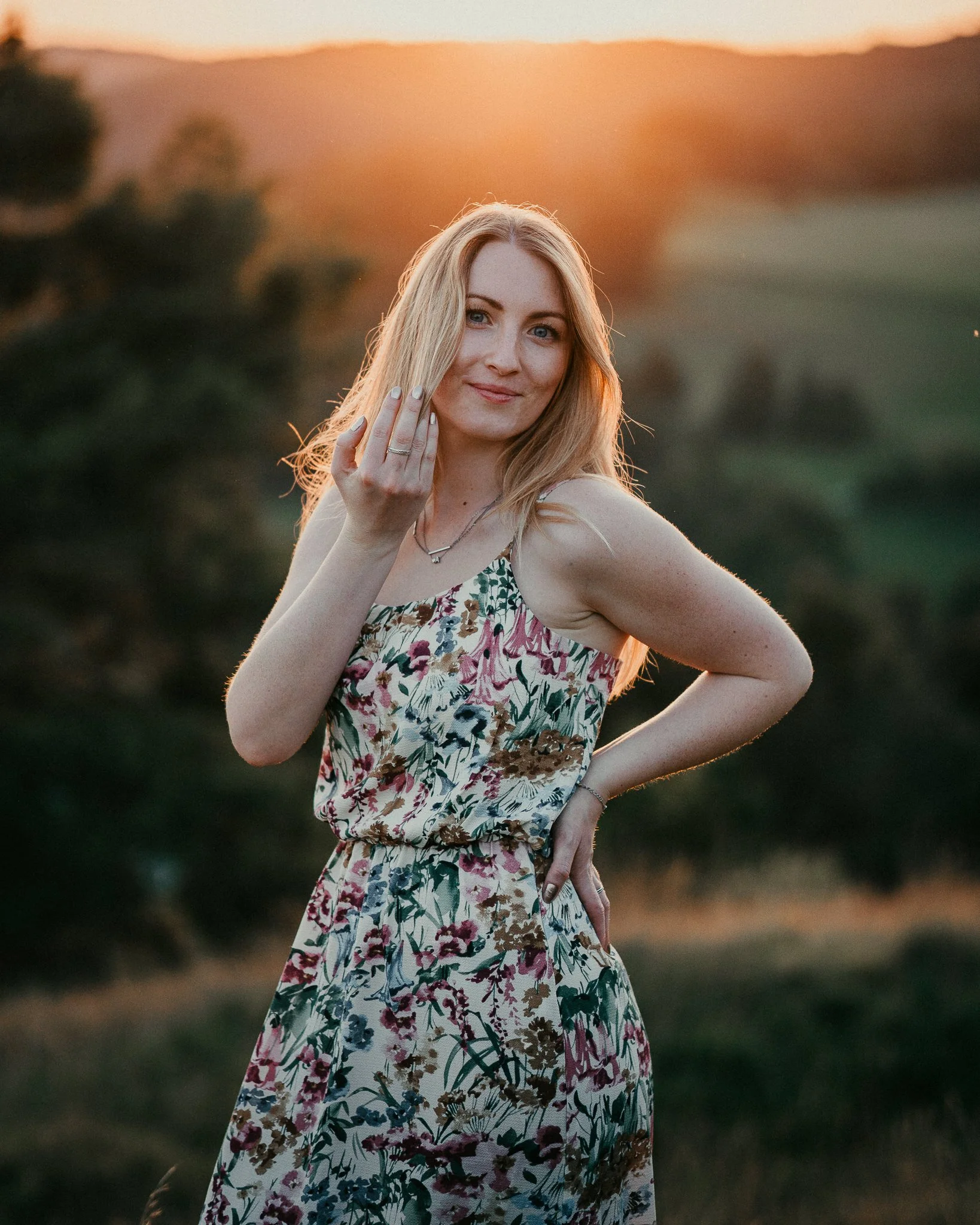 Eine Frau in einem Blumenmantel steht im Freien bei Sonnenuntergang, die Frau lächelt sanft und hat die Hand am Gesicht. Sie trägt eine Halskette und einen Ring.