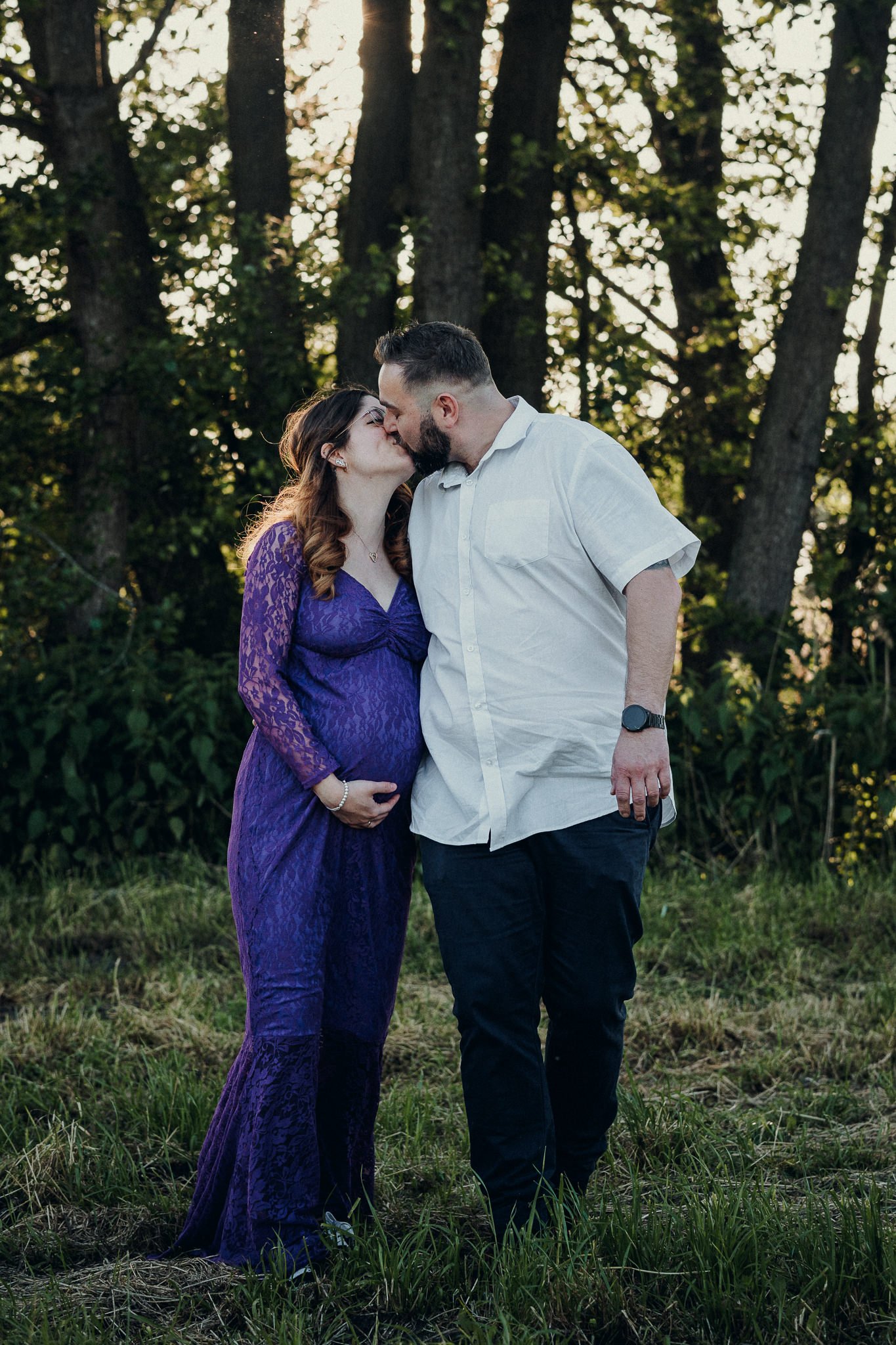 Ein Paar, eine schwangere Frau in lila Kleid und ein Mann, küssen sich in einem Wald bei Sonnenuntergang.