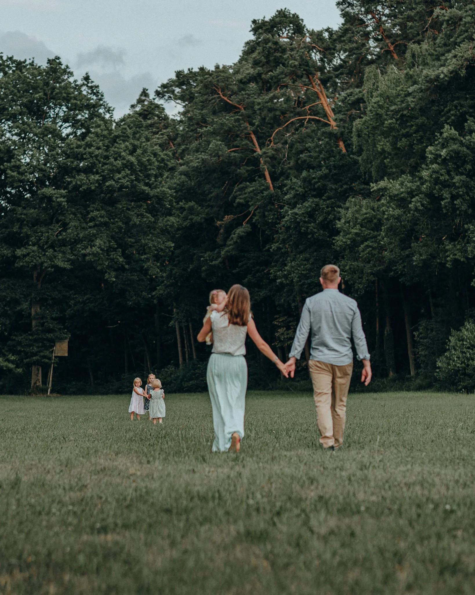 Familie mit zwei Erwachsenen und drei Kindern beim Spaziergang auf einer Wiese vor einem Wald.