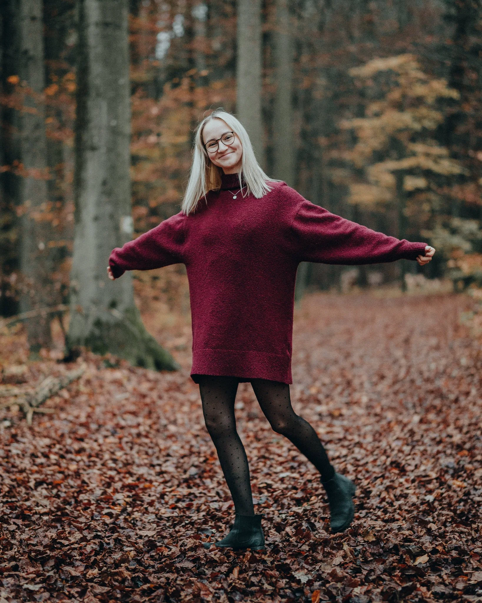 Eine lachende junge Frau mit Brille trägt einen roten Pullover und schwarze Strümpfe mit Punkten, steht auf einem Laubweg im Herbstwald.