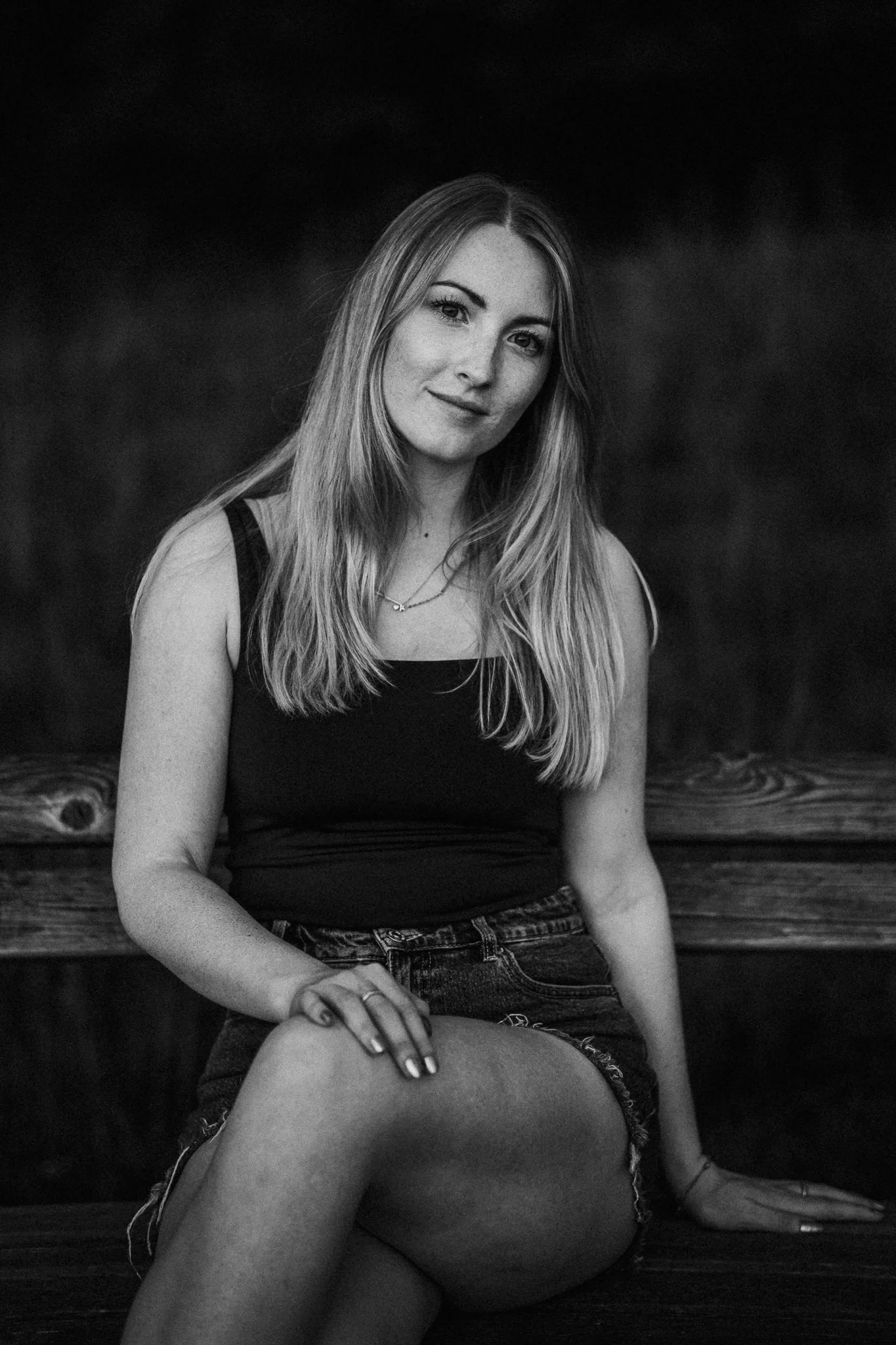 Schwarz-Weiß-Fotografie einer jungen Frau mit langen, geraden Haaren, sitzend, im dunklen Hintergrund.