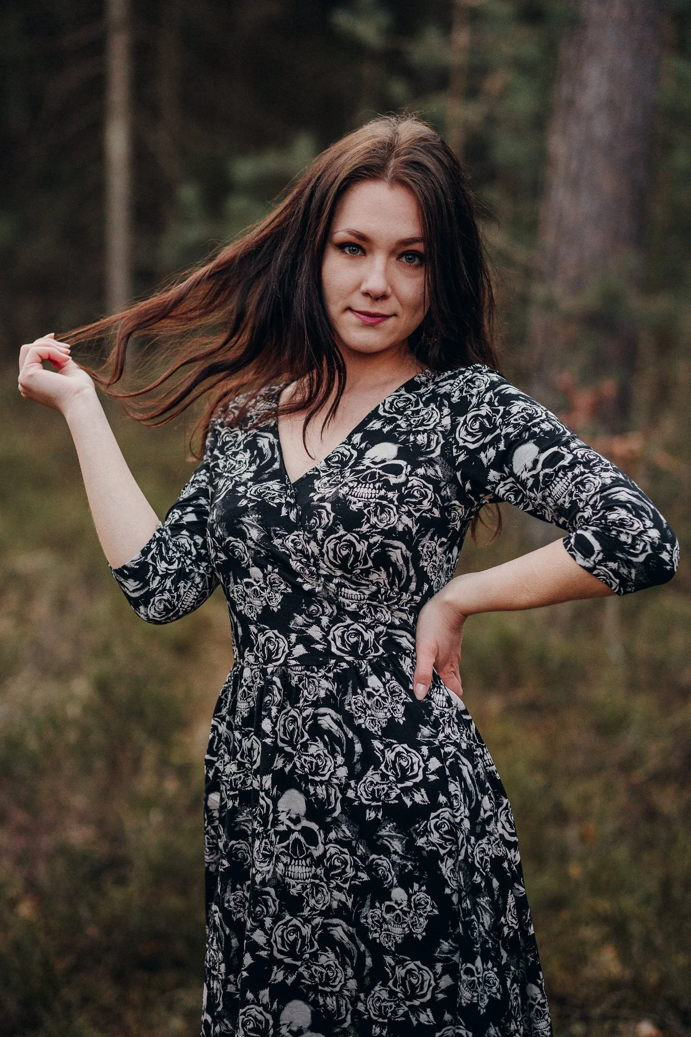 Eine junge Frau mit braunen Haaren und blauen Augen steht in einem Wald und trägt ein schwarzes Kleid mit weißen Rosen- und Totenkopfdesigns.